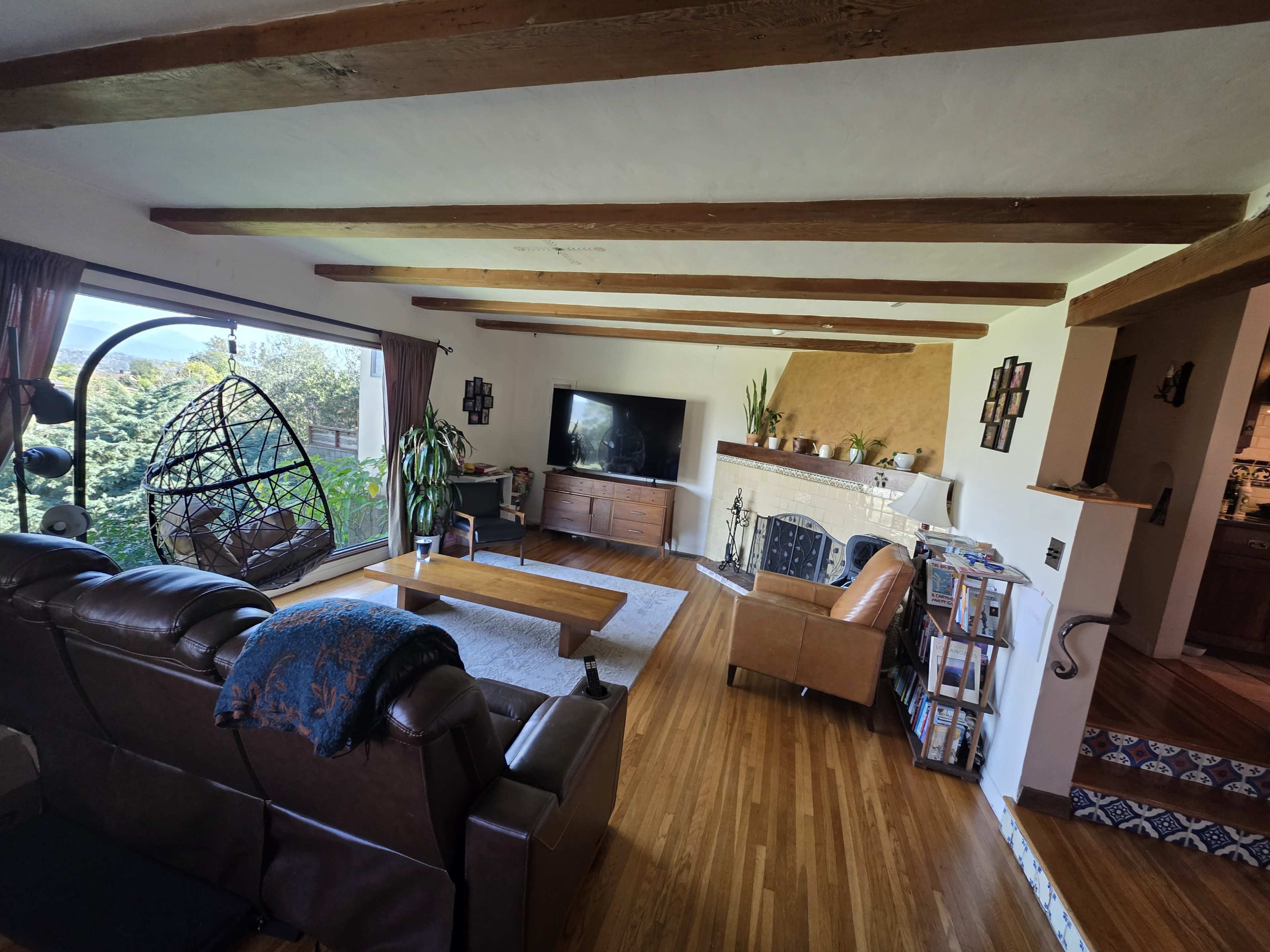 The image shows a living room with a large window, a hanging chair, a television, wooden beams on the ceiling, and a mix of furniture arranged around a coffee table.