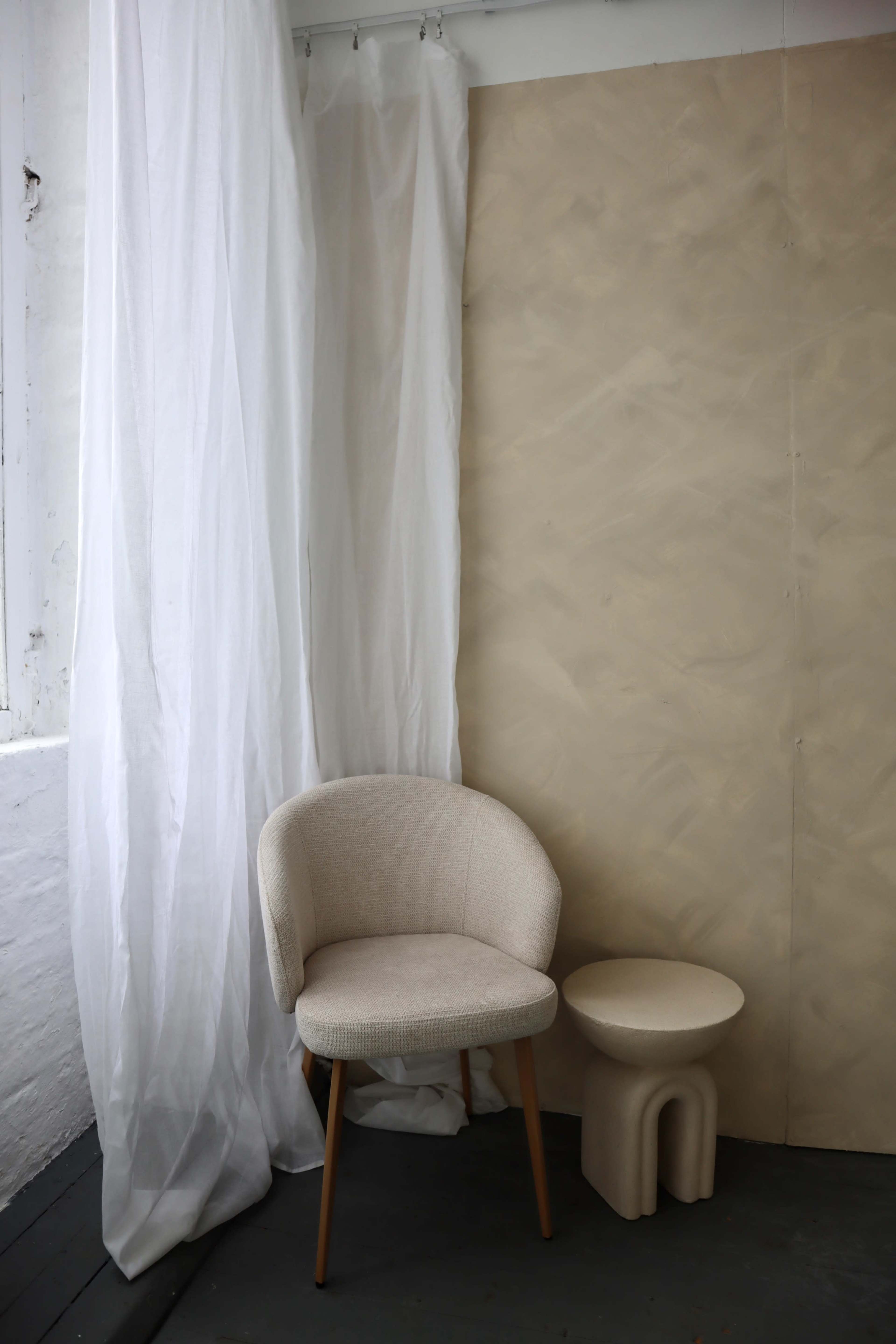 A light-colored chair is positioned beside a small, rounded stool in a minimalist interior setting with a white curtain and a textured beige wall.