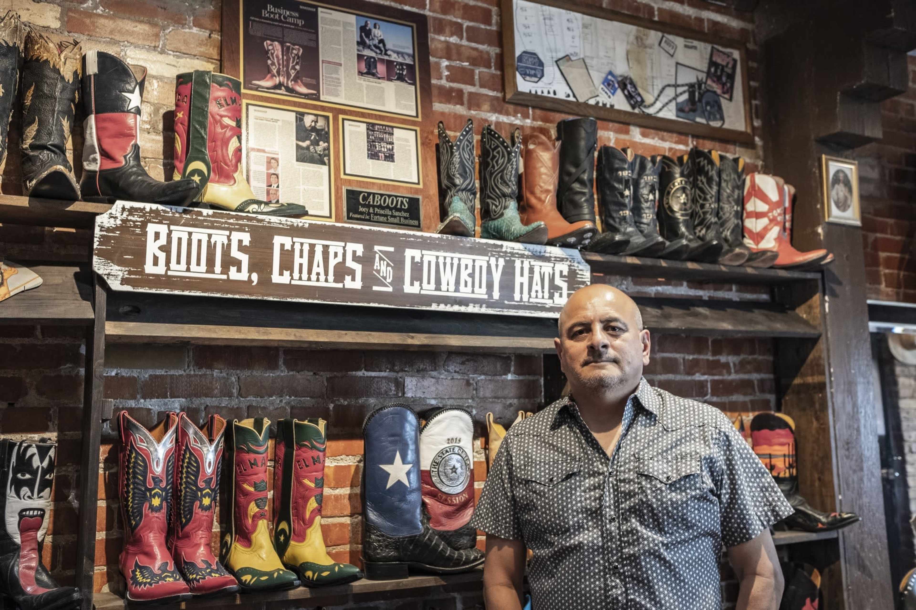 A man stands in front of a display shelf featuring a variety of colorful cowboy boots, with a sign that reads "BOOTS, CHAPS & COWBOY HATS" above.
