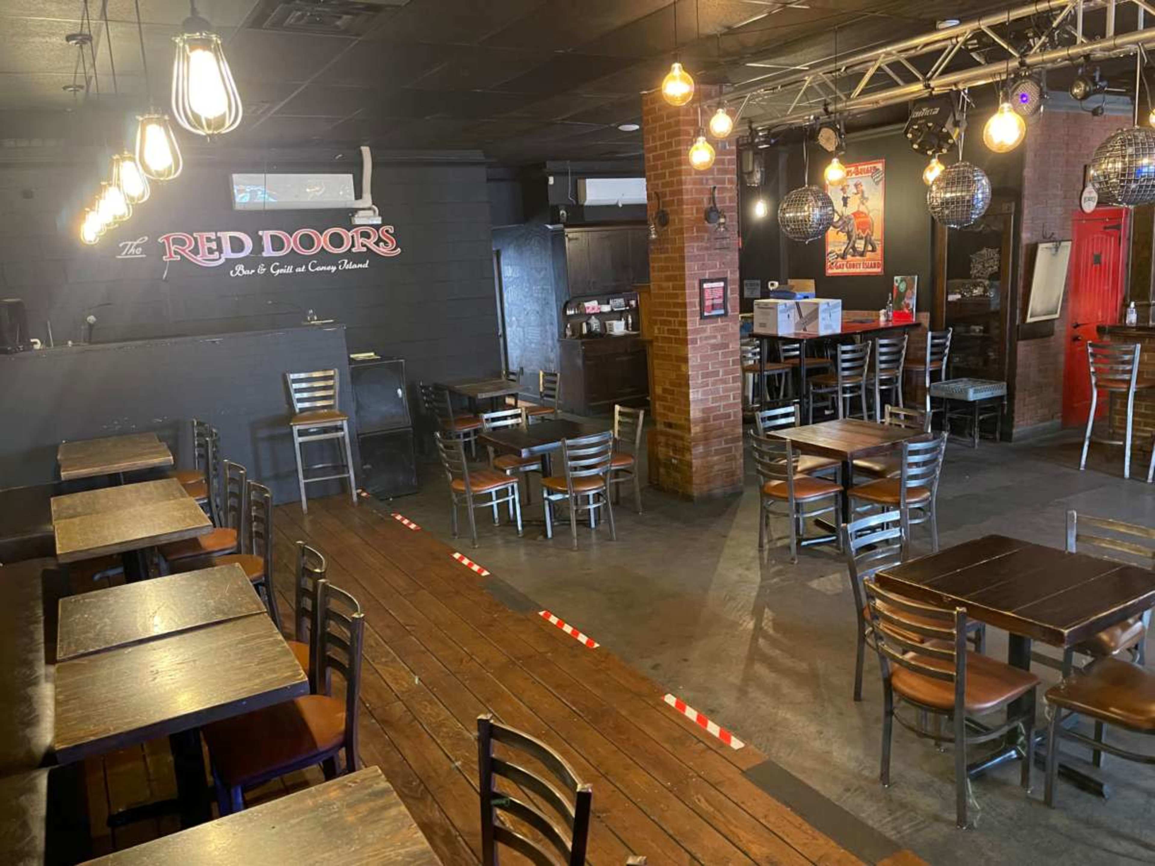 The image shows an interior view of a restaurant or bar featuring wooden tables and chairs, pendant lighting, and a stage area.