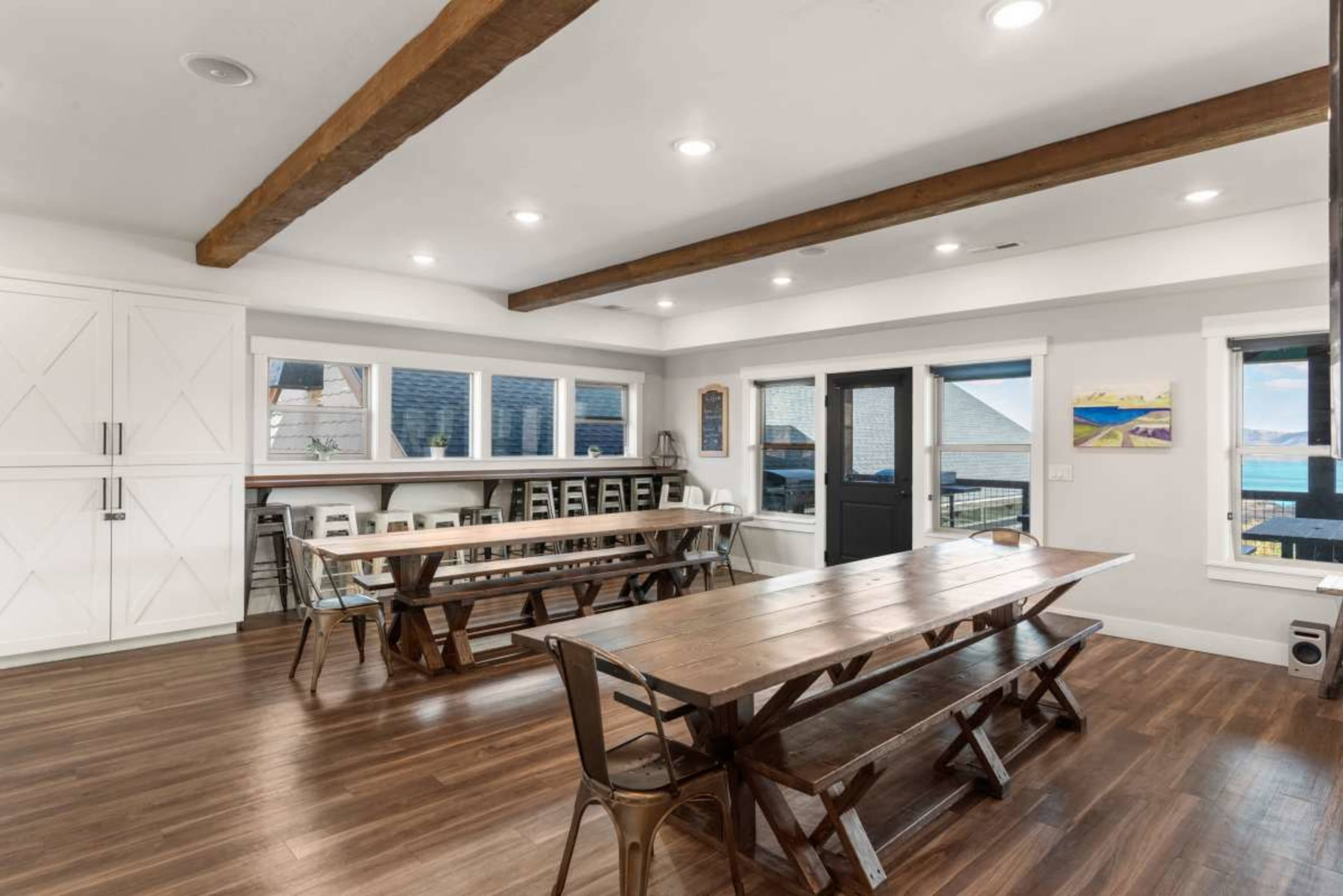 A spacious dining area features long wooden tables with metal chairs, large windows, and exposed wooden beams on the ceiling.