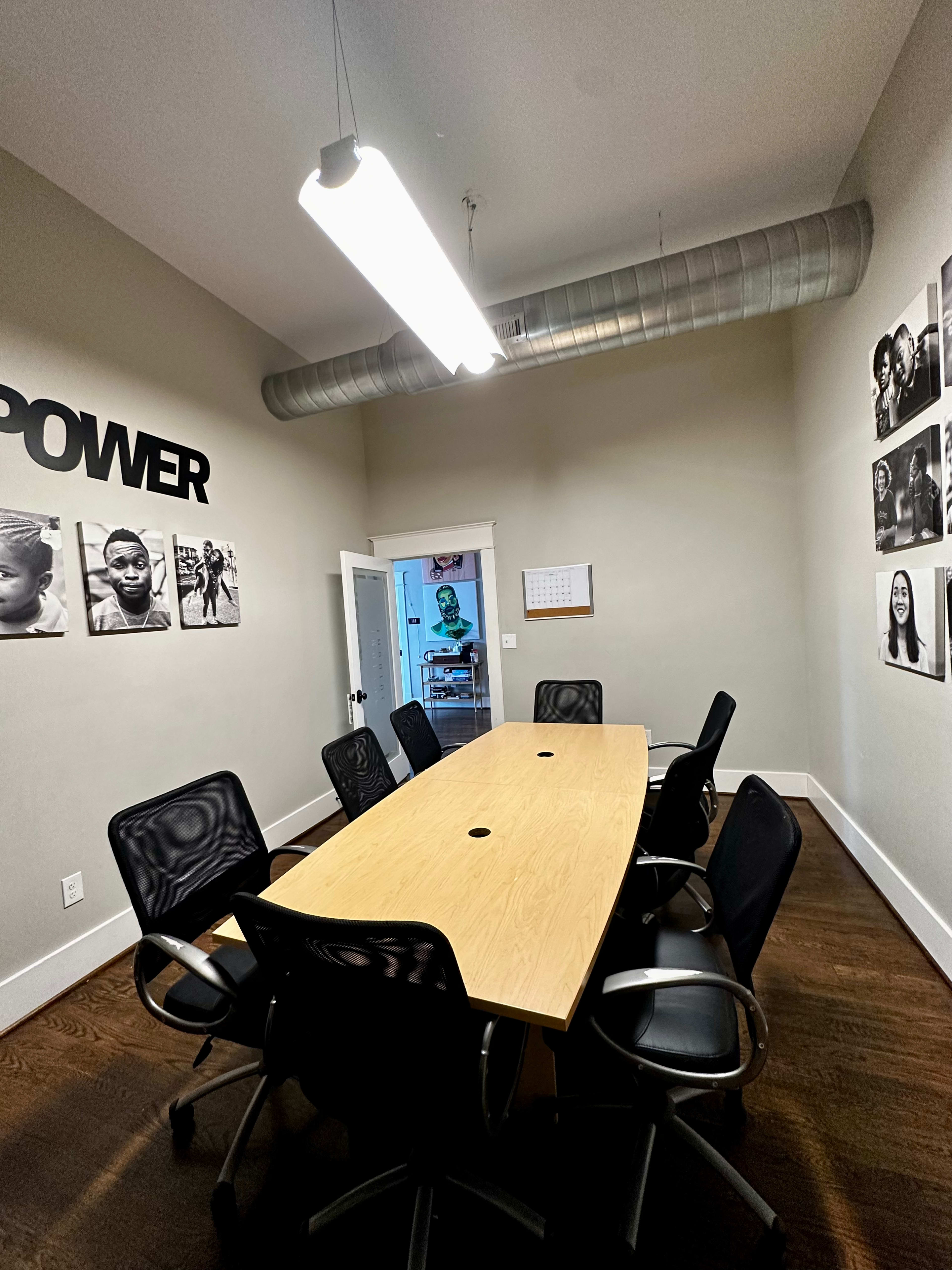 A conference room features a long wooden table surrounded by black mesh chairs, with black-and-white photographs on the walls and a door leading to another room in the background.