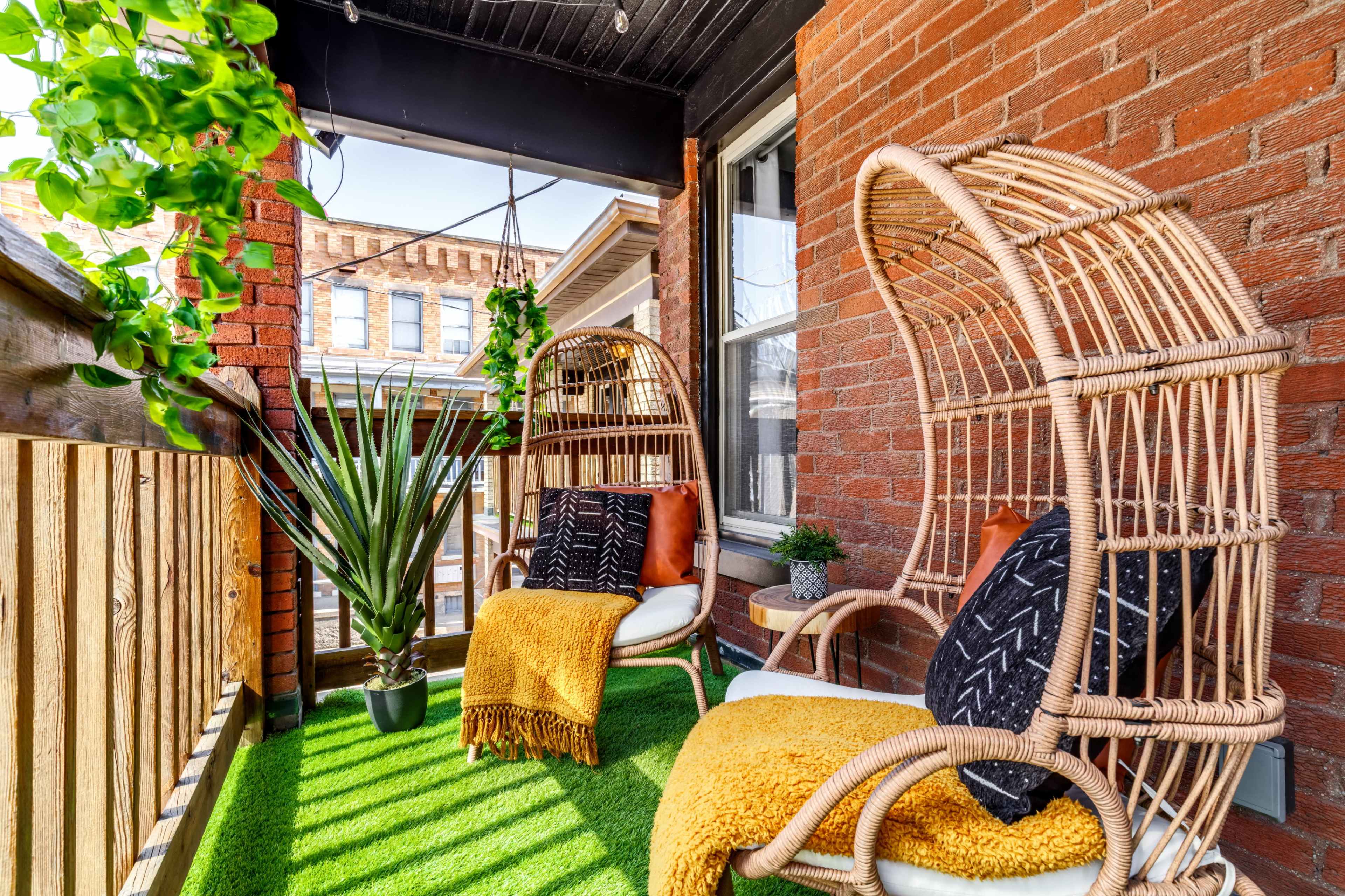A balcony features two wicker chairs with cushions, surrounded by potted plants and artificial grass.