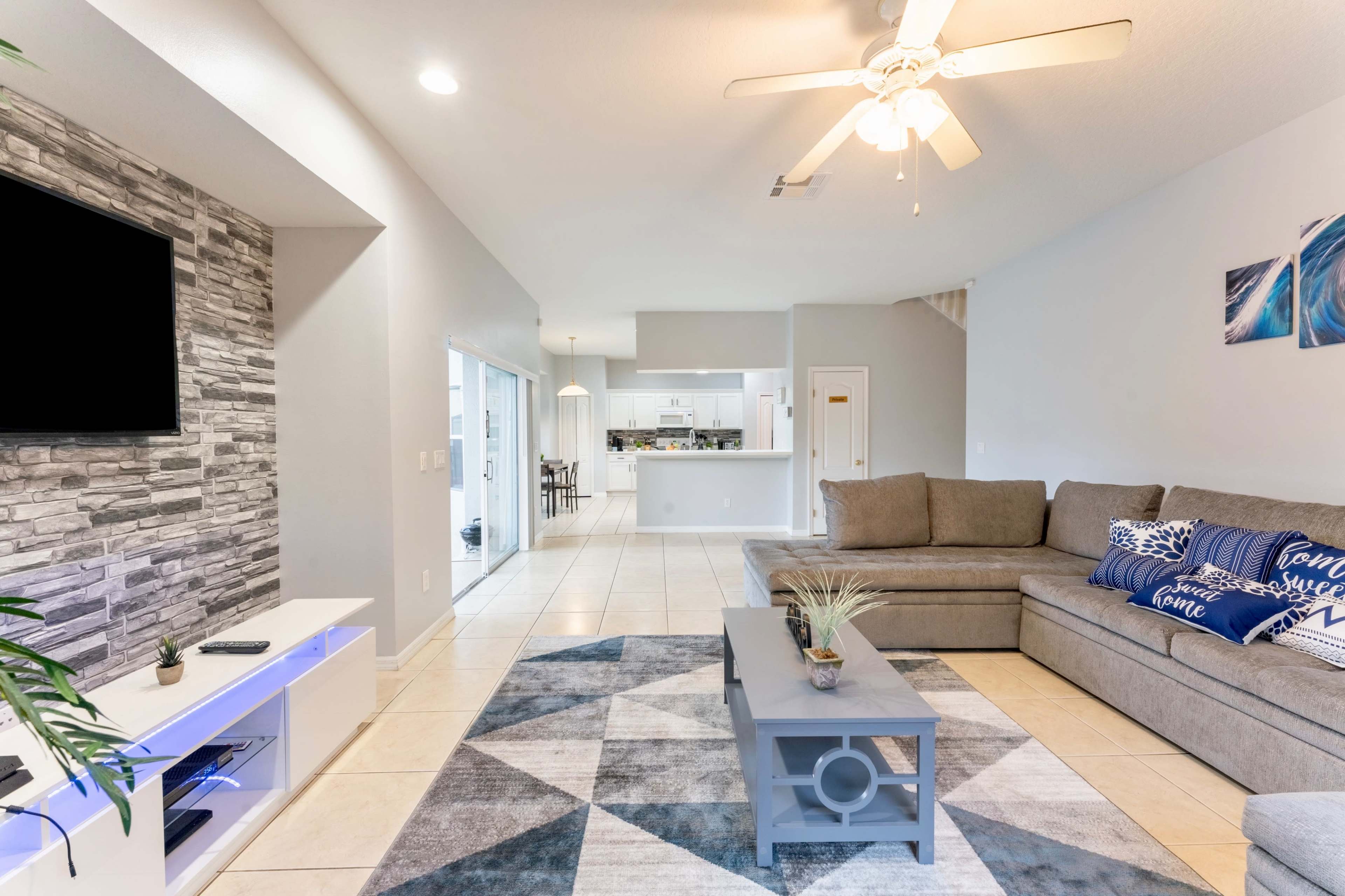 The living room features a large sectional sofa, a coffee table, and a TV mounted on a stone accent wall, with an open view to a dining area behind it.