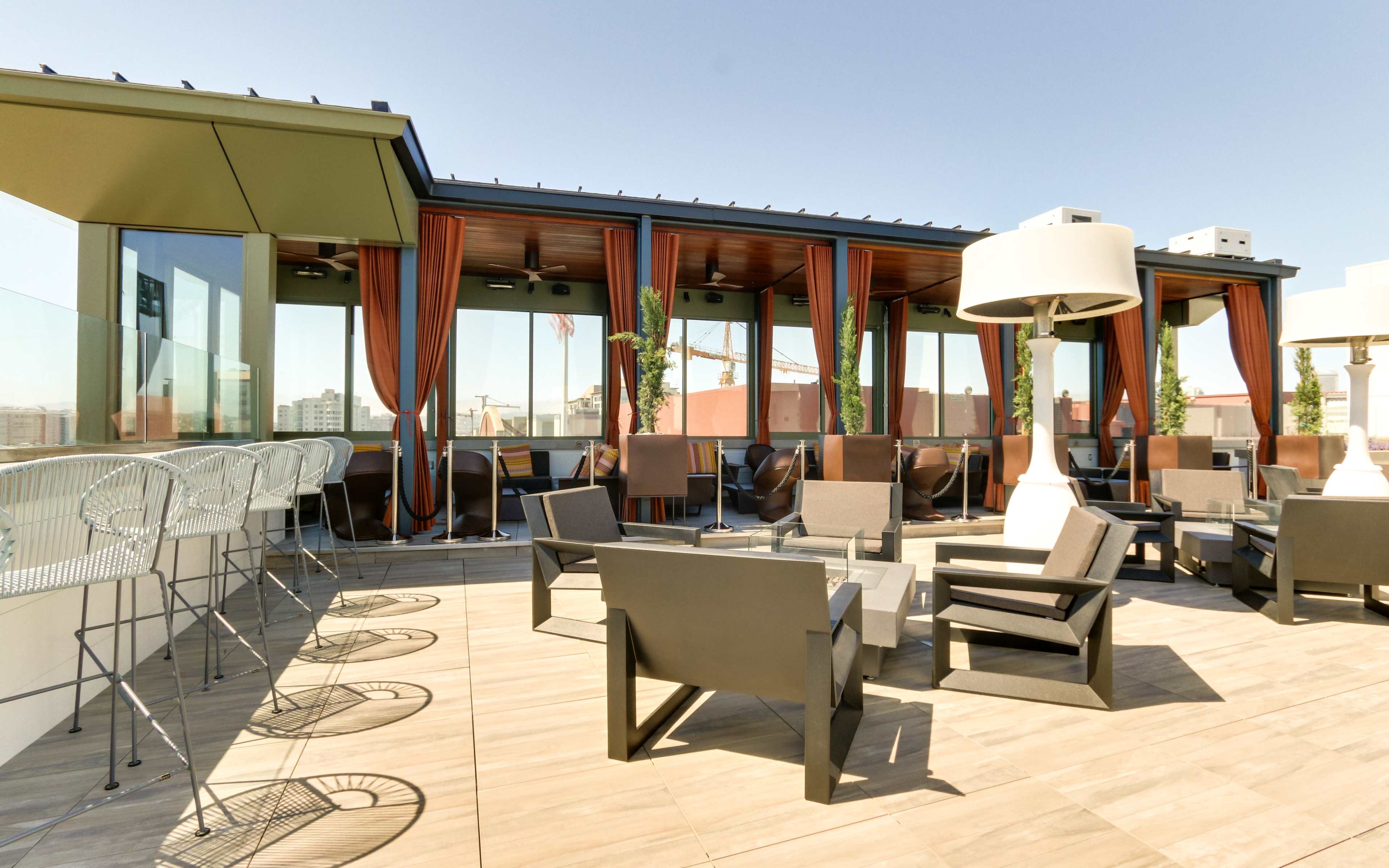 A rooftop lounge area with modern seating arrangements, glass barriers, and a bar counter under clear blue skies.