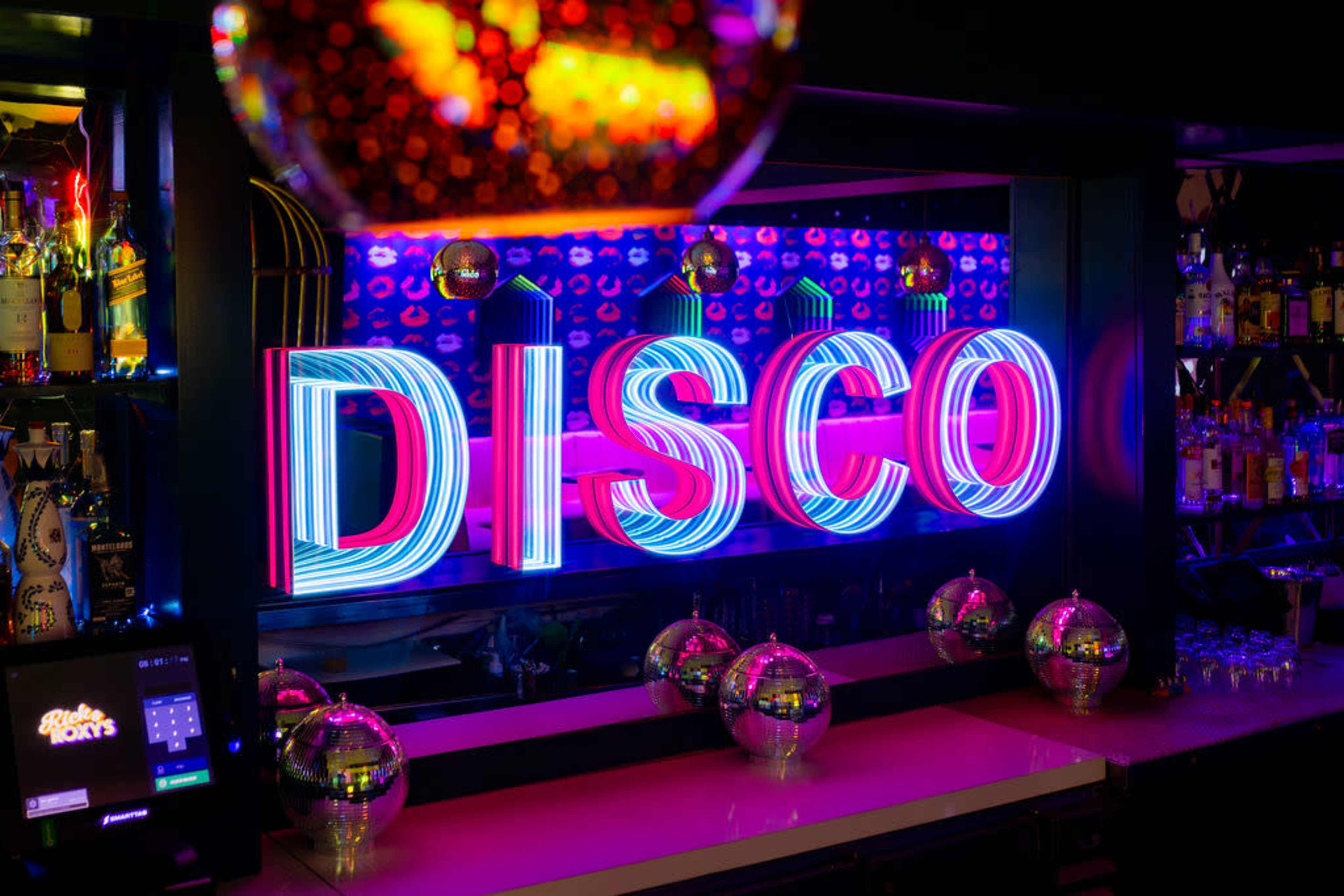 The image features a brightly lit "DISCO" sign in neon colors, with mirrored disco balls placed on a counter in a bar setting.