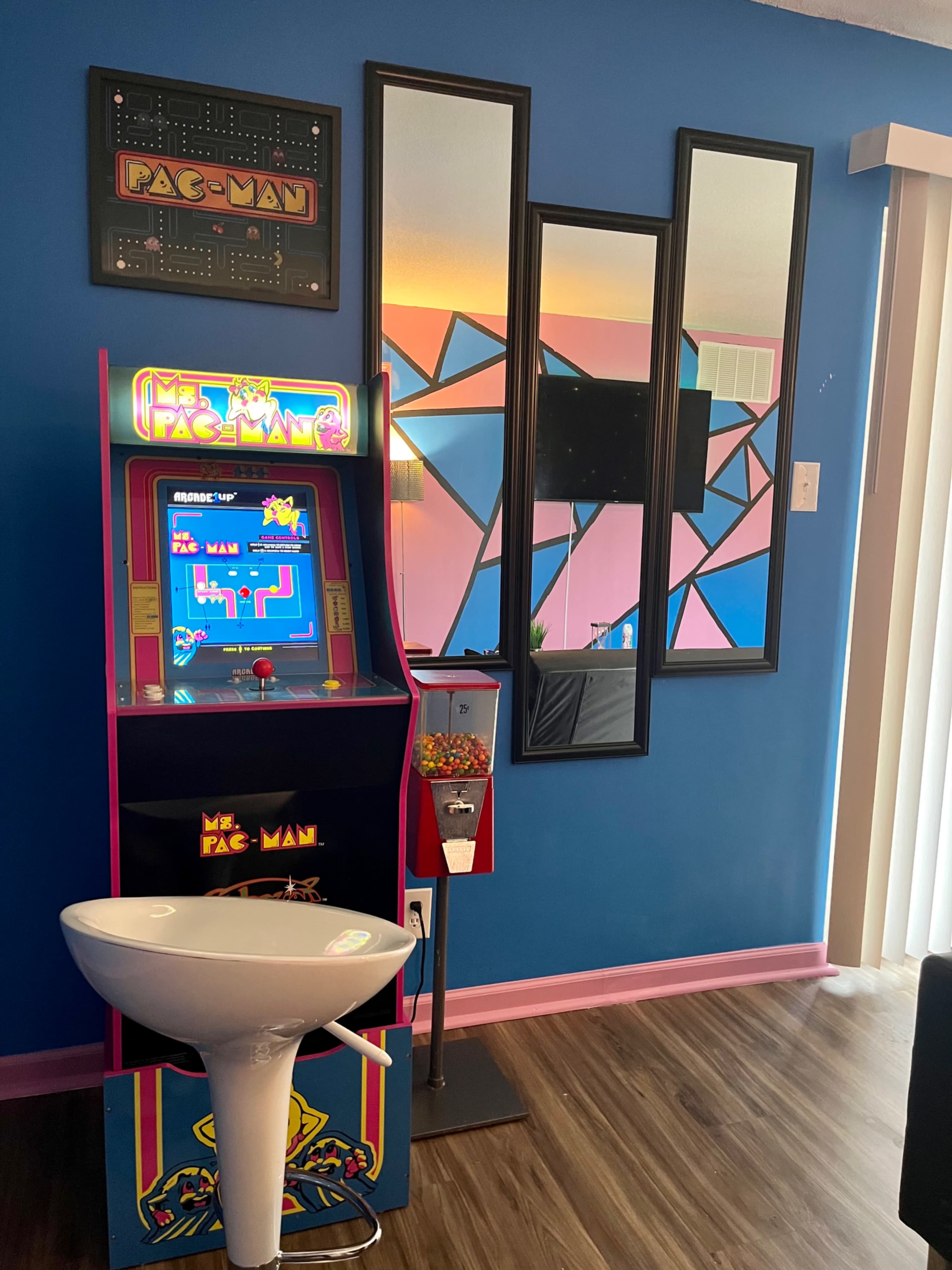 A retro Pac-Man arcade machine stands next to a modern sink in a brightly colored room with patterned mirrors and a television.