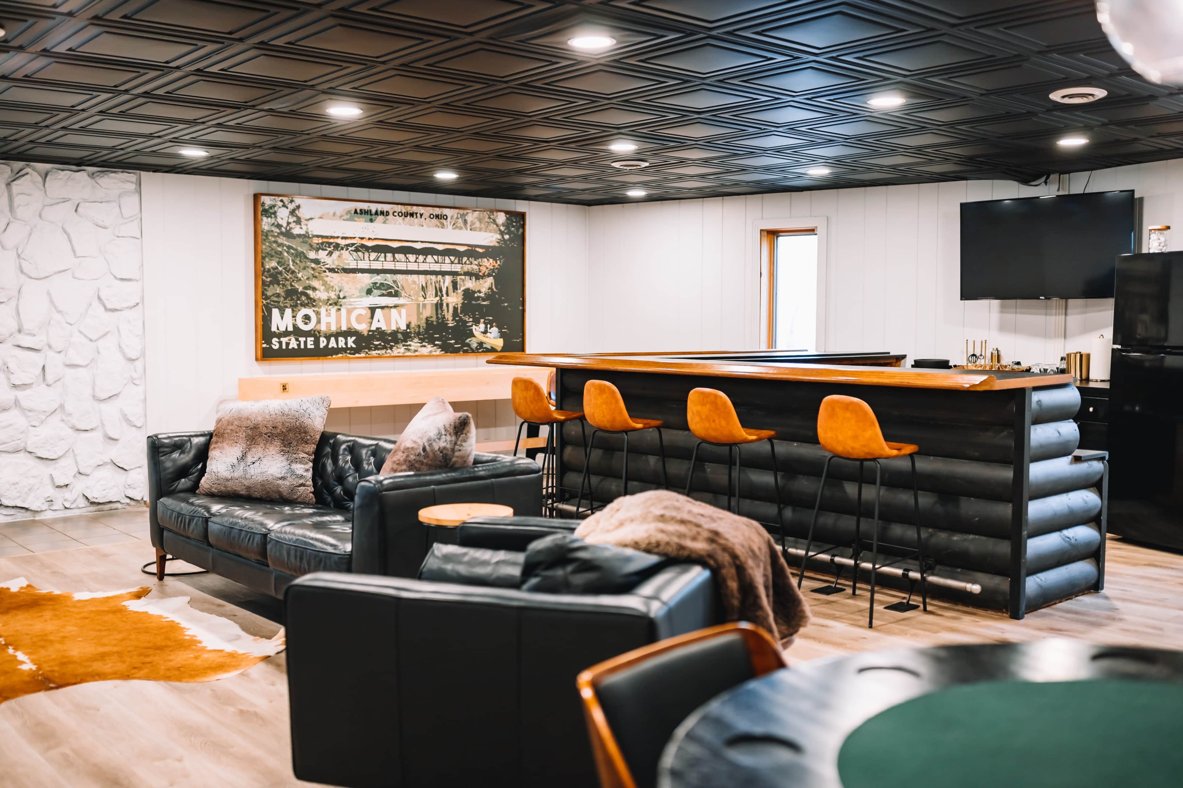The image shows a modern interior living space with a bar area, leather seating, and a large wall-mounted television, complemented by a decorative backdrop featuring Mohican State Park.