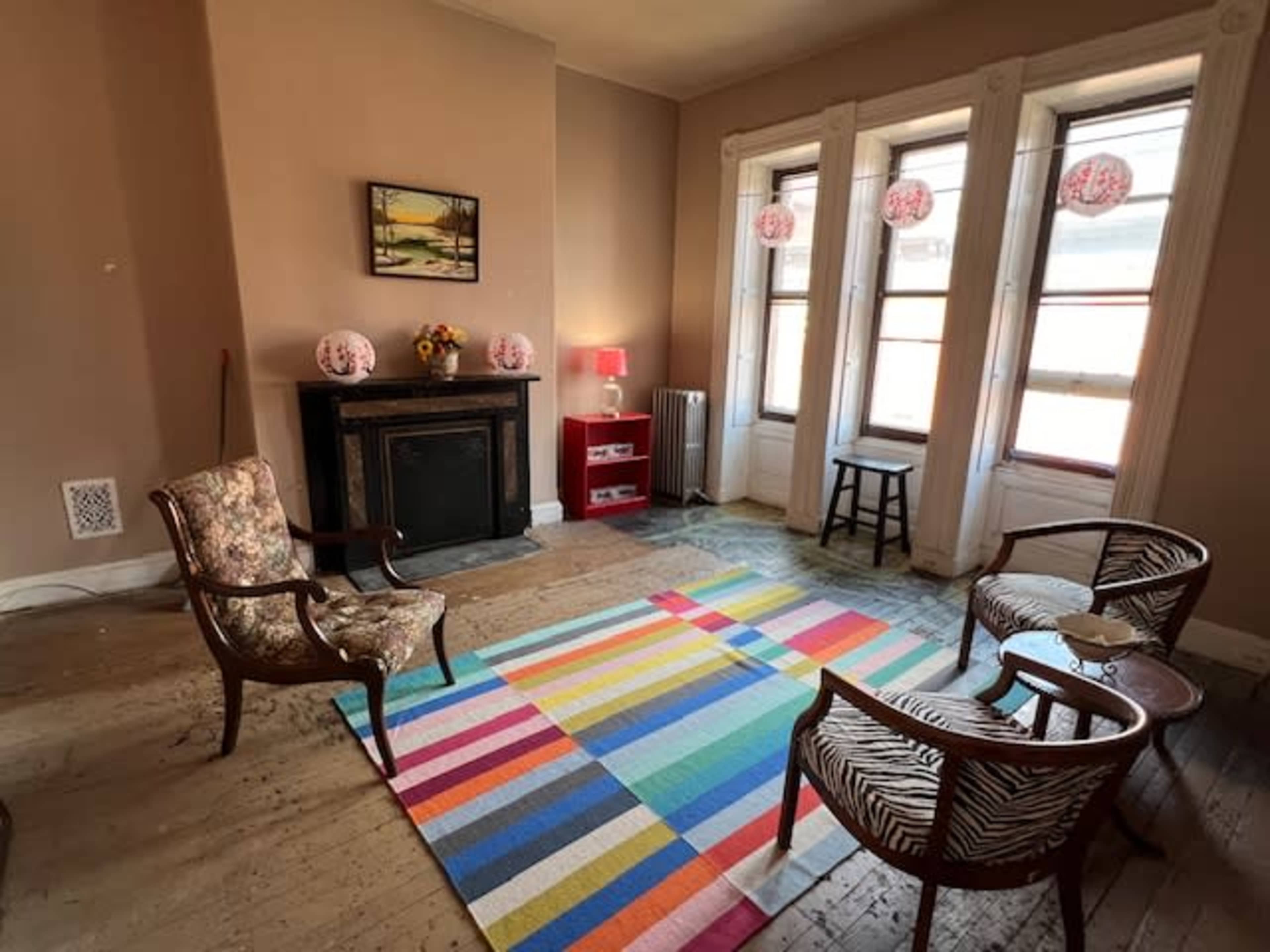 A living room with a striped rug, two chairs, a small red table, and a fireplace, along with three windows letting in natural light.