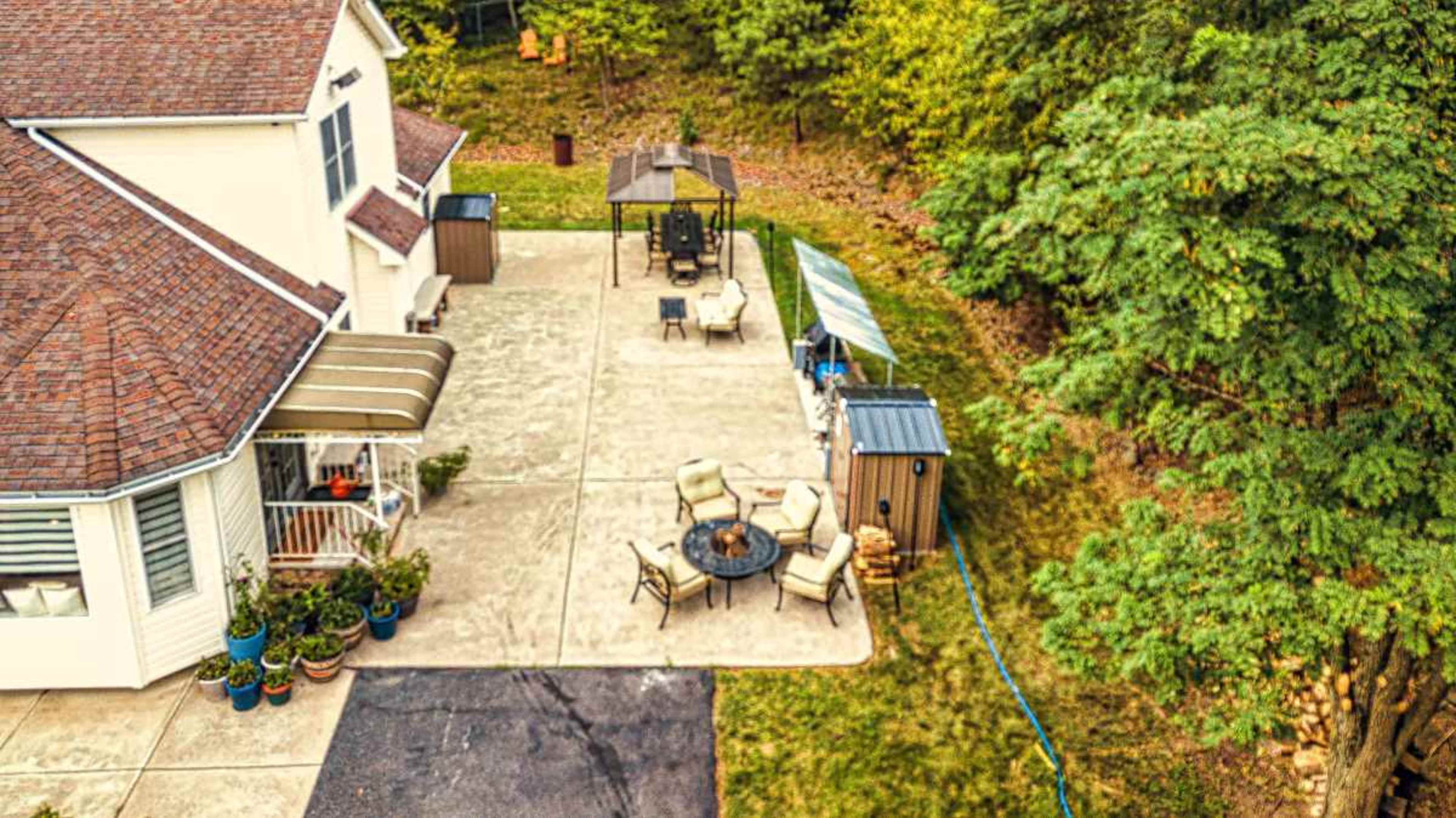 The image shows a backyard patio with seating, a fire pit, and a small shed beside a house surrounded by trees.
