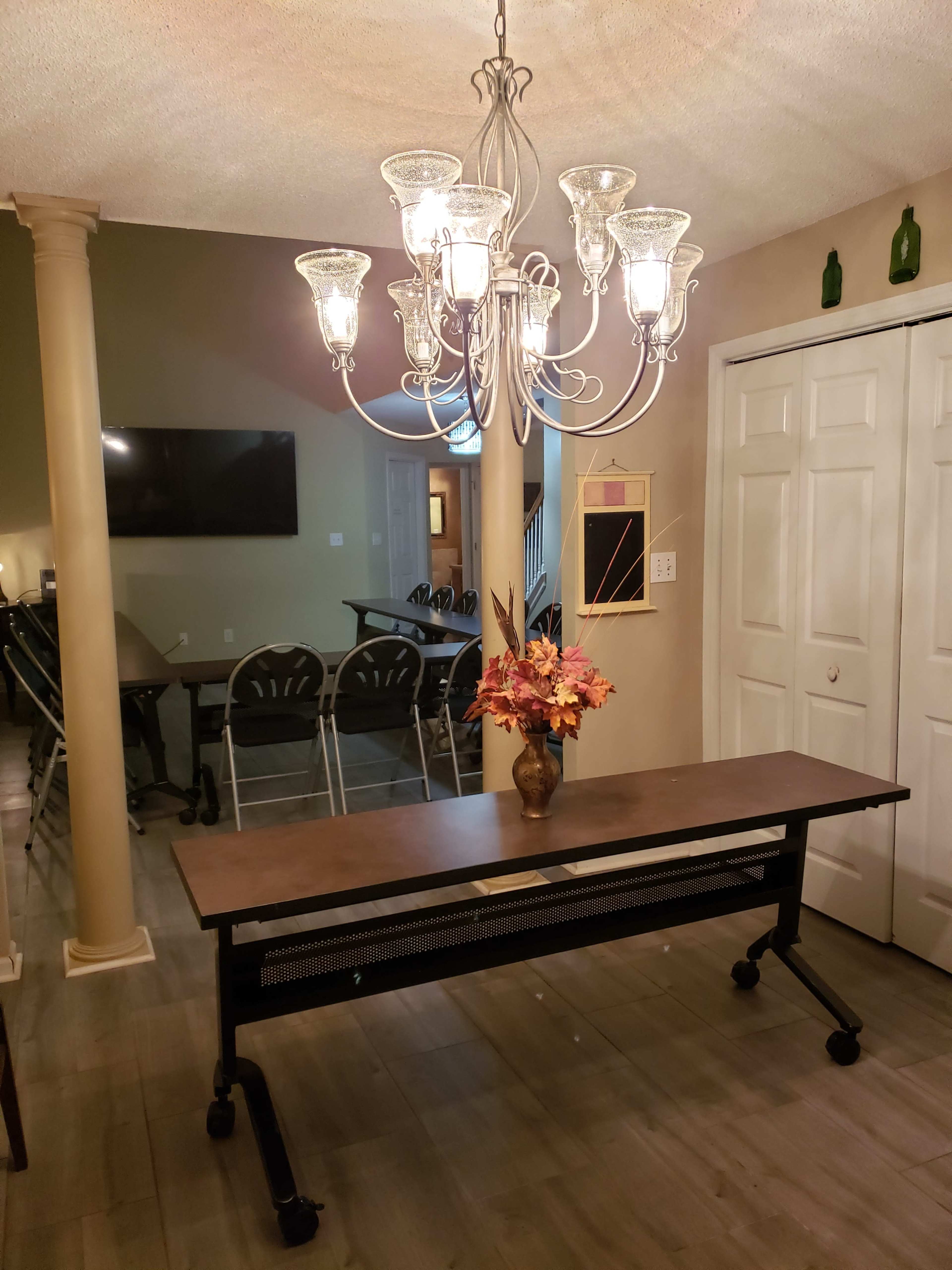 A chandelier hangs above a table with a flower vase in a room that features a mounted TV and folding chairs.