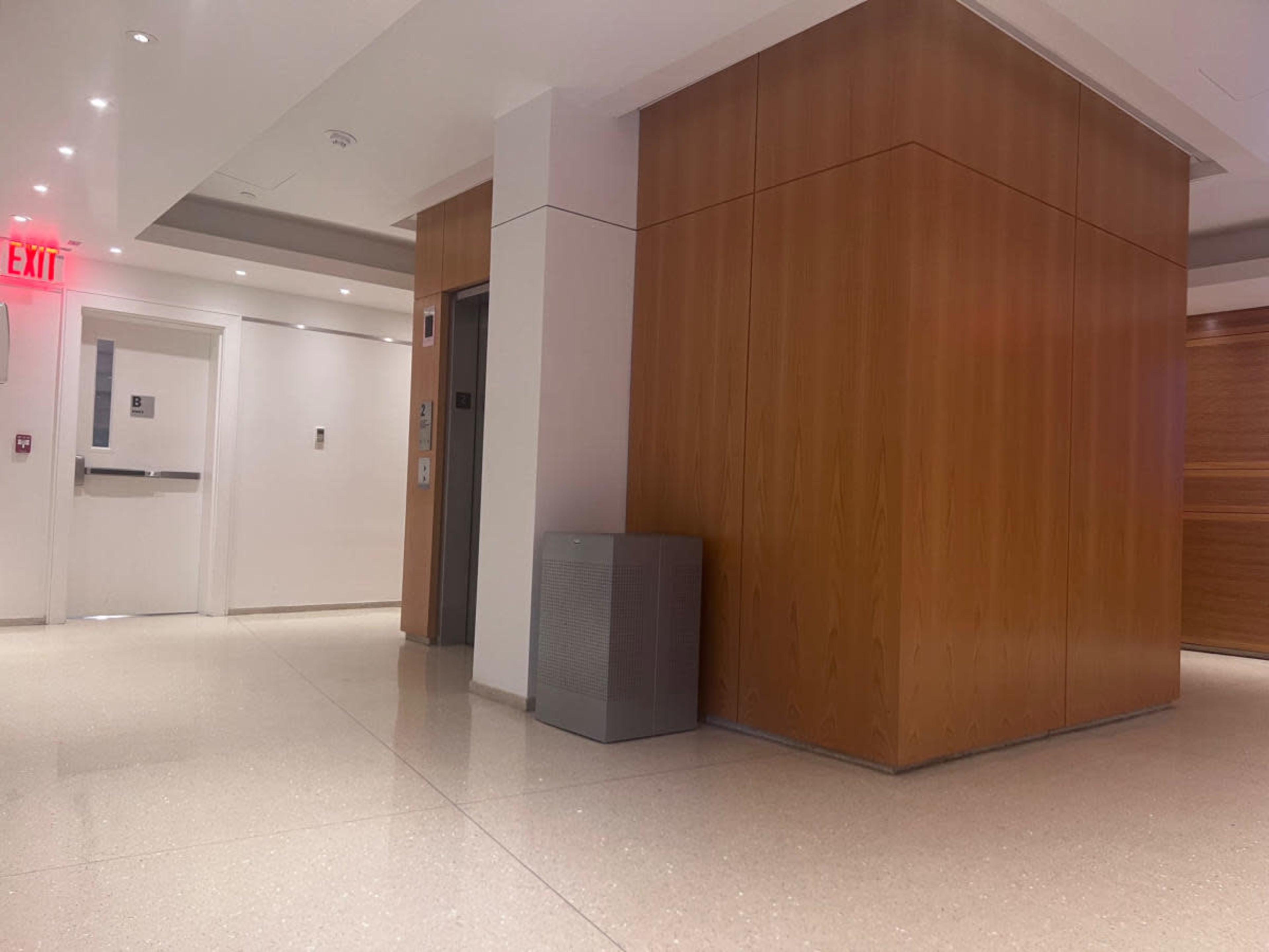 The image shows a modern lobby area featuring an elevator, a doorway, and wooden wall paneling.