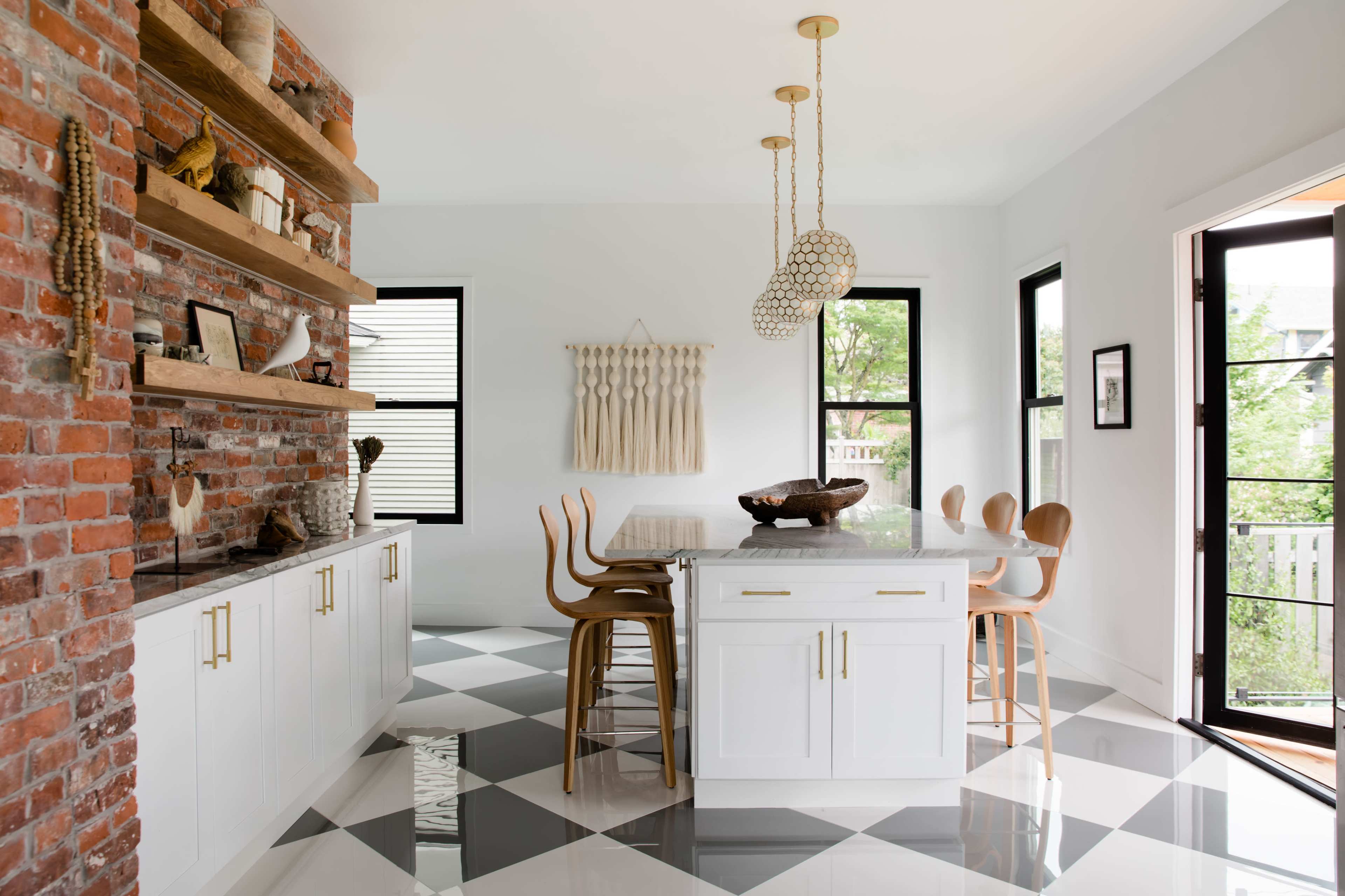 A modern kitchen features a brick wall, open shelving with decorative items, a central island with a wooden bowl, and a dining table surrounded by wooden chairs on a checkered floor.