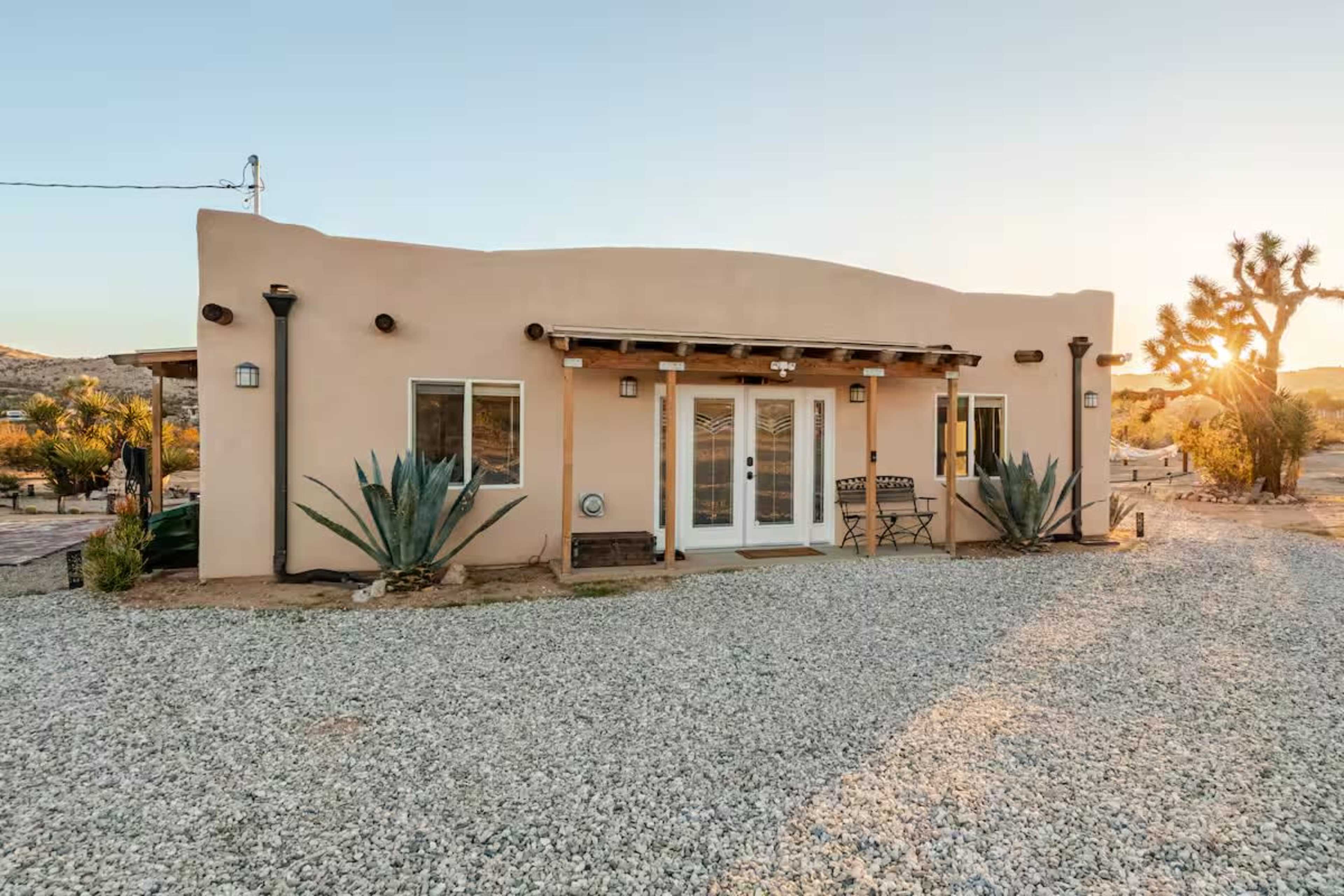 A single-story adobe-style house with large front windows is situated in a gravel landscape surrounded by desert vegetation.