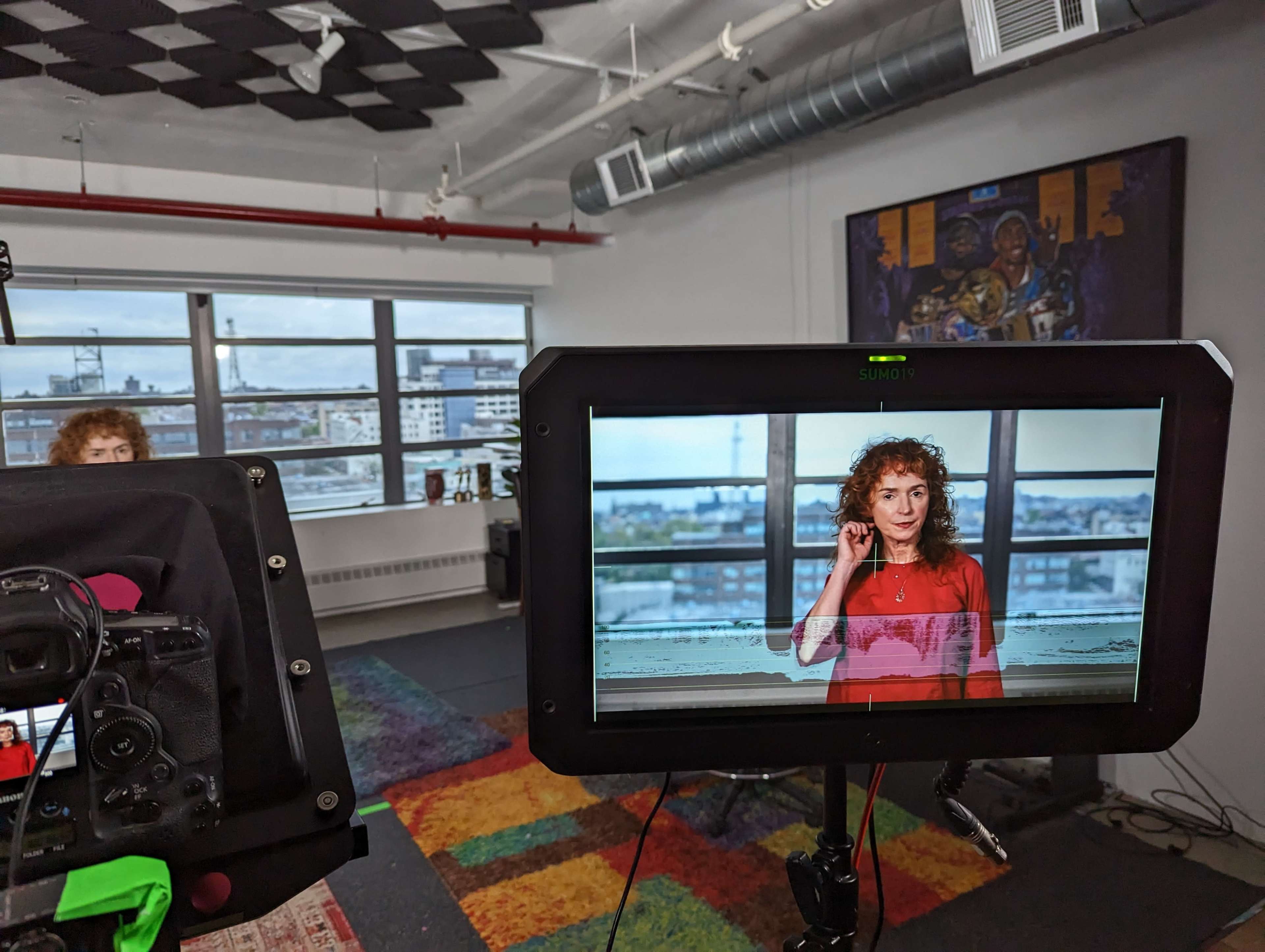 A woman with curly red hair stands in front of a large window in a brightly lit studio, while a camera set up displays her on a monitor.