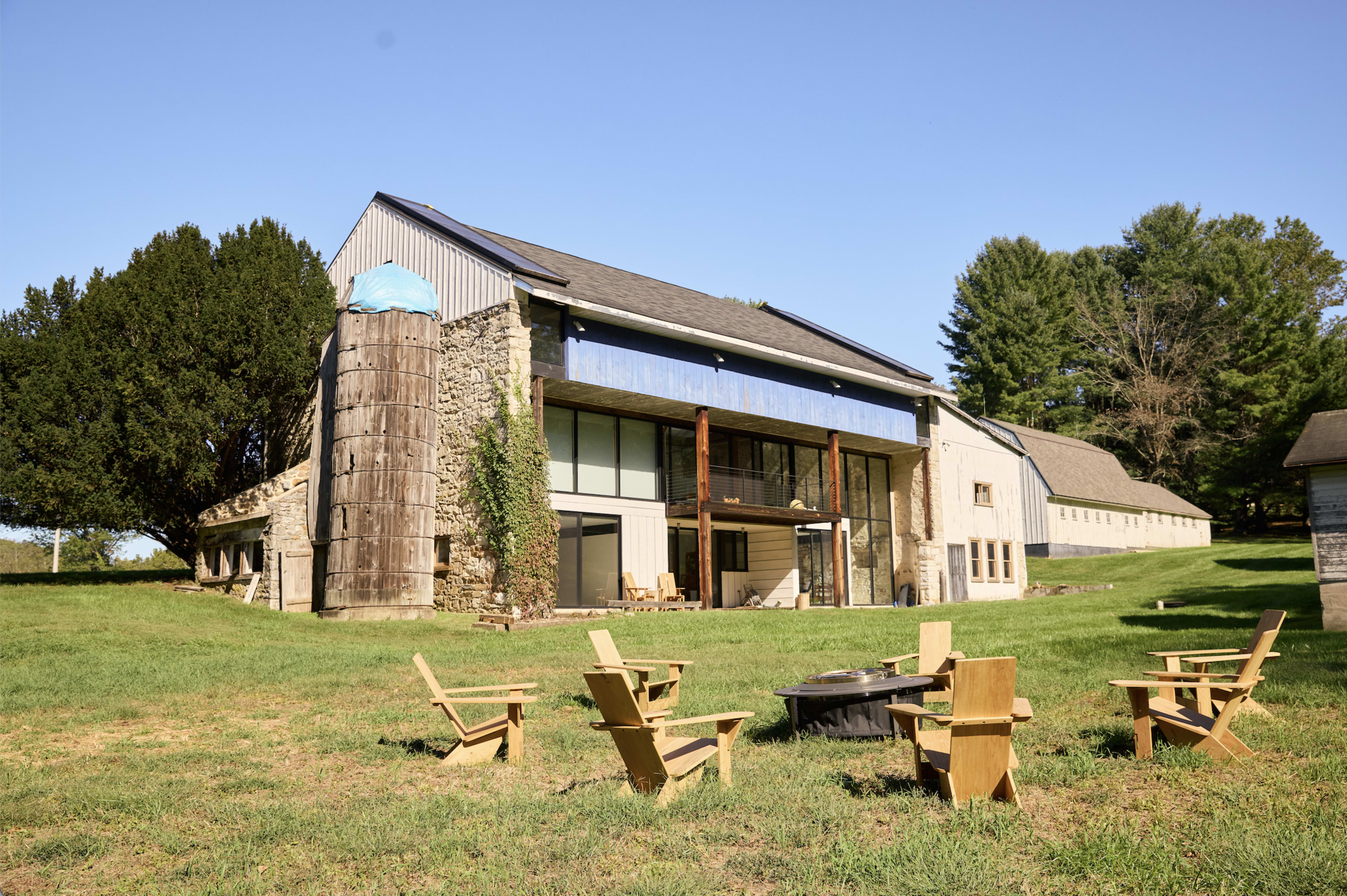 A modern building with large windows and a stone structure is situated on a green lawn, surrounded by trees, and features a circular seating arrangement made of wooden chairs and a fire pit in the foreground.