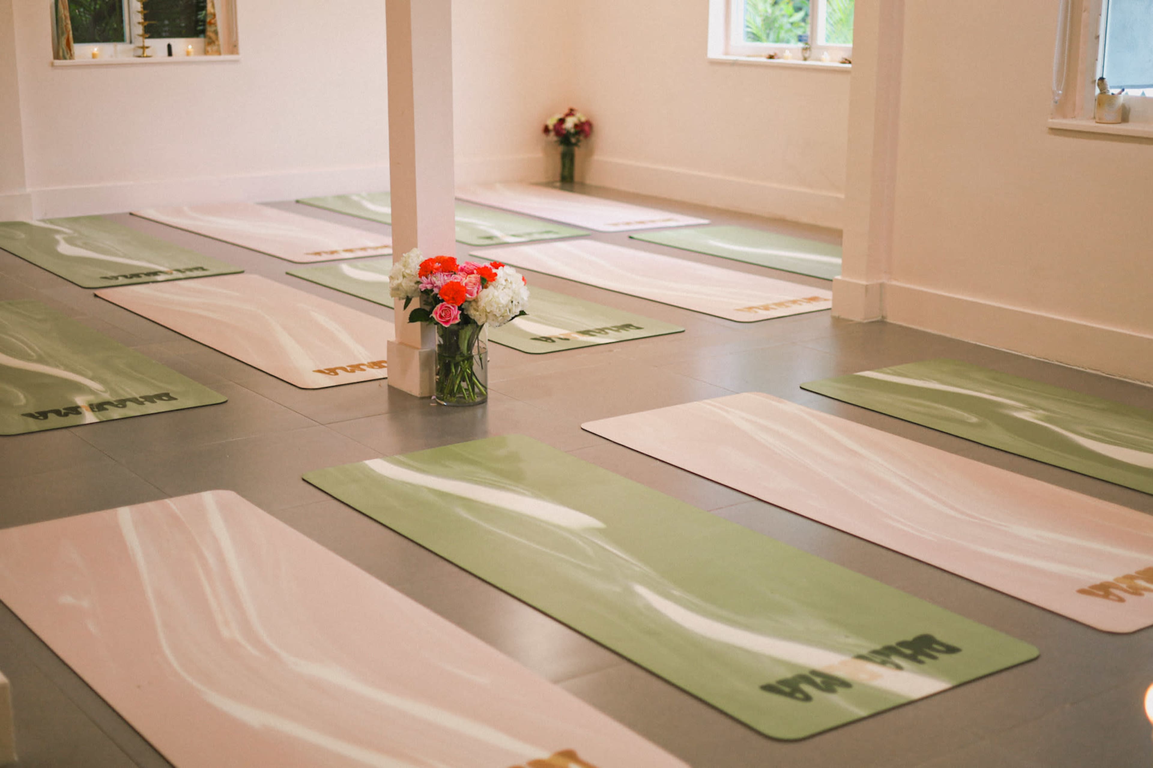 The image shows a yoga studio with multiple pastel-colored mats arranged neatly on a gray floor, and a vase of flowers in the center.