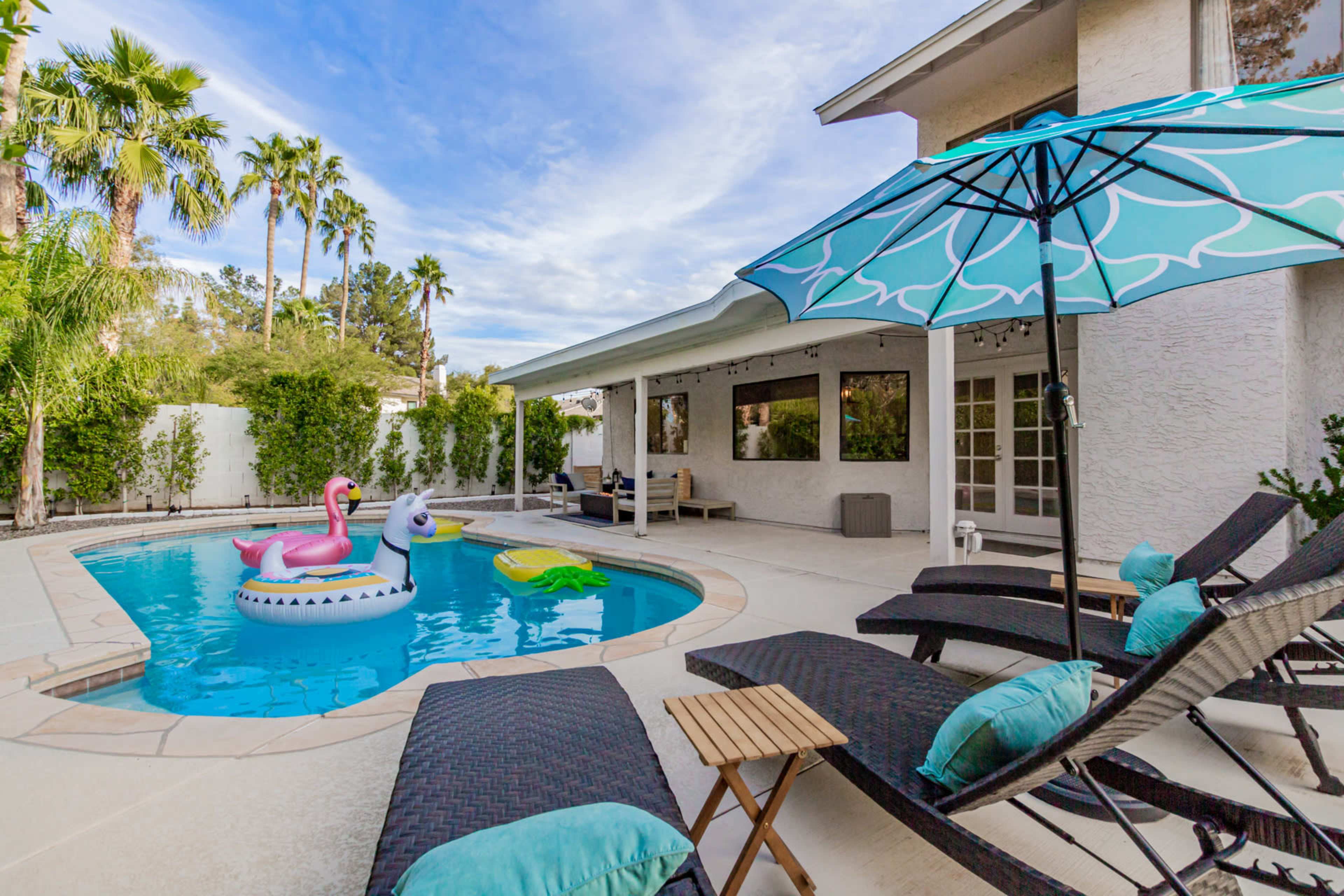 A backyard pool area featuring lounge chairs, a large umbrella, and inflatable pool toys in the water.