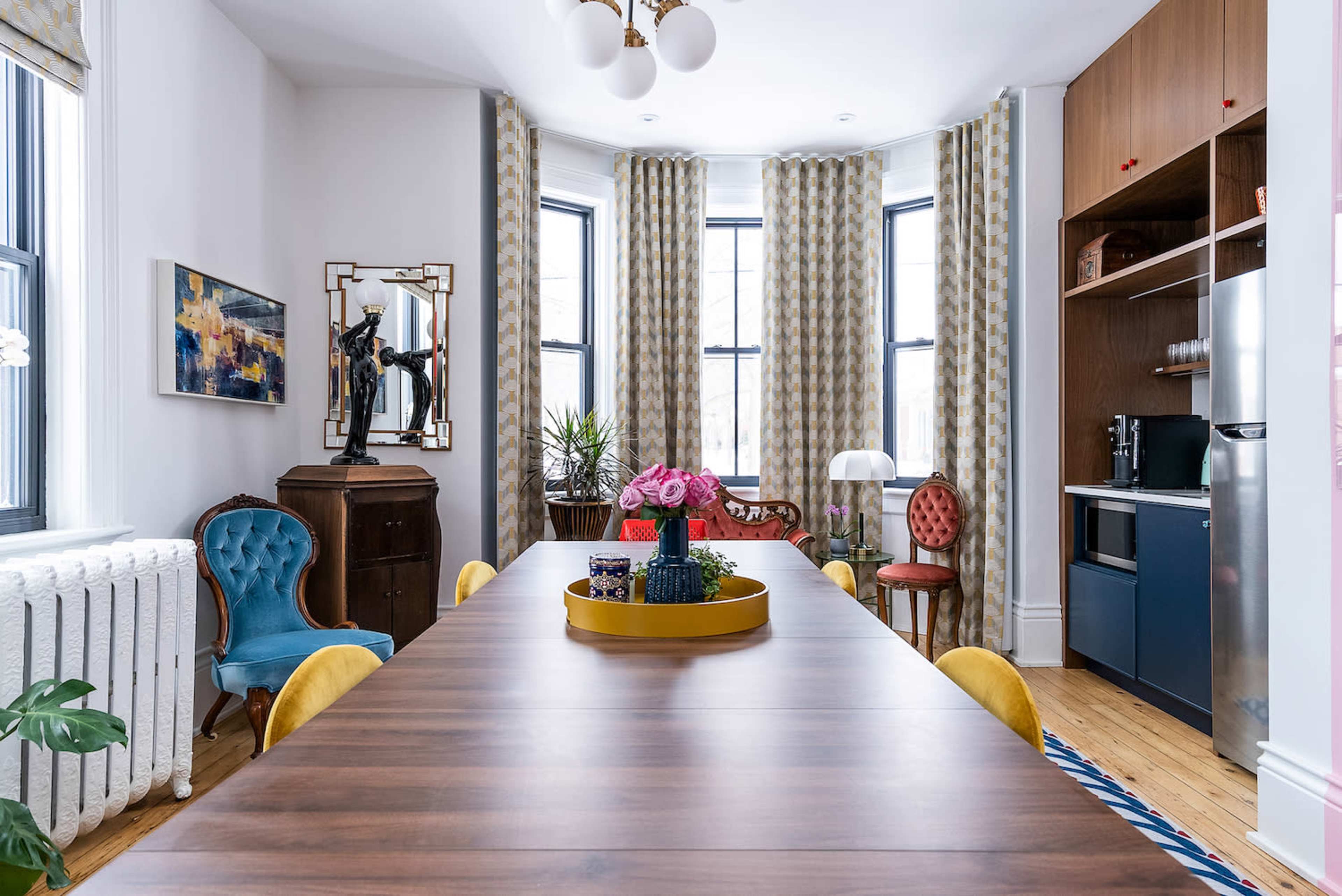 A dining area features a long wooden table surrounded by colorful chairs, with a bay window, curtains, and a kitchen cabinetry in the background.
