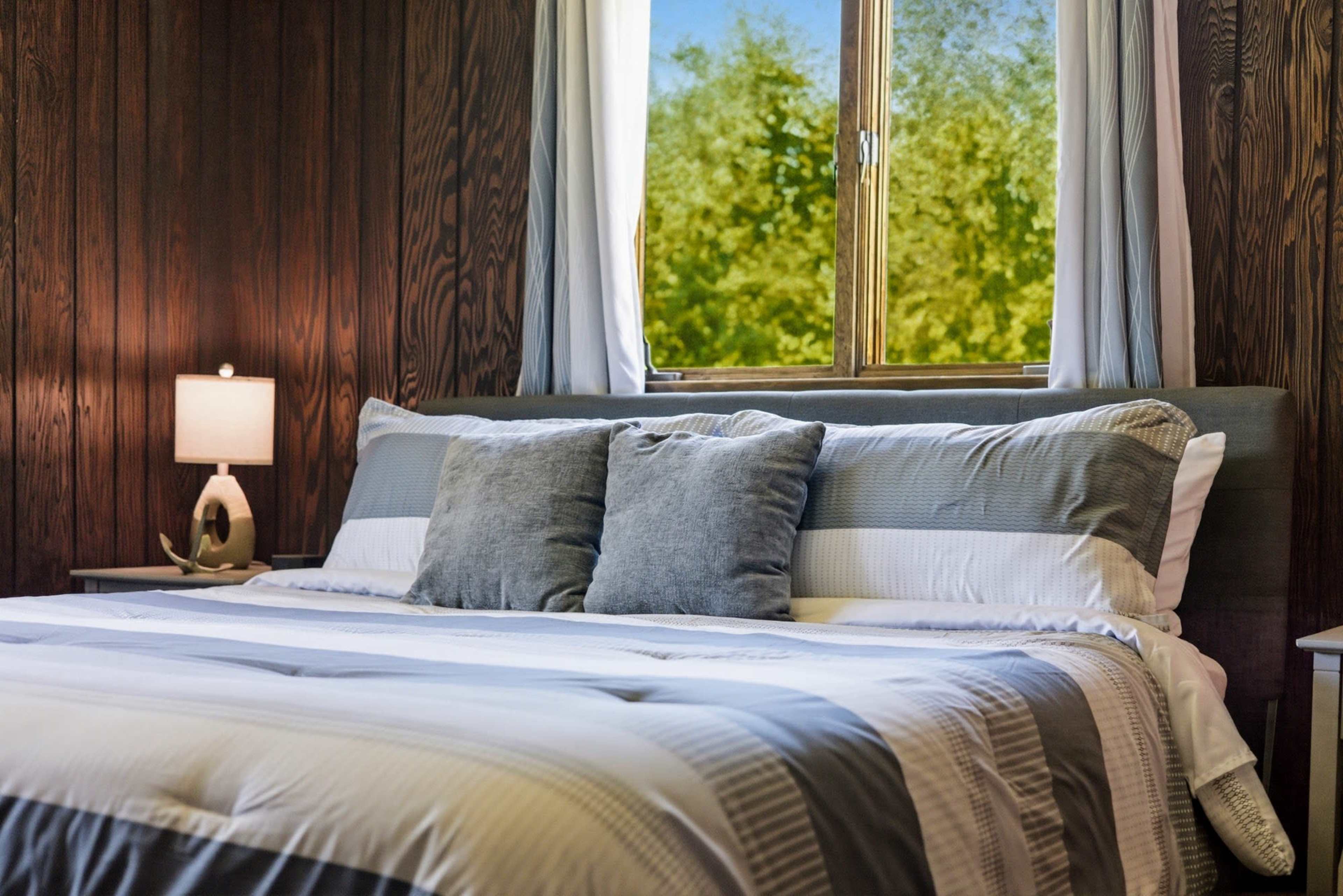 The image shows a neatly made bed with gray and white bedding, two decorative pillows, and a window revealing greenery outside.
