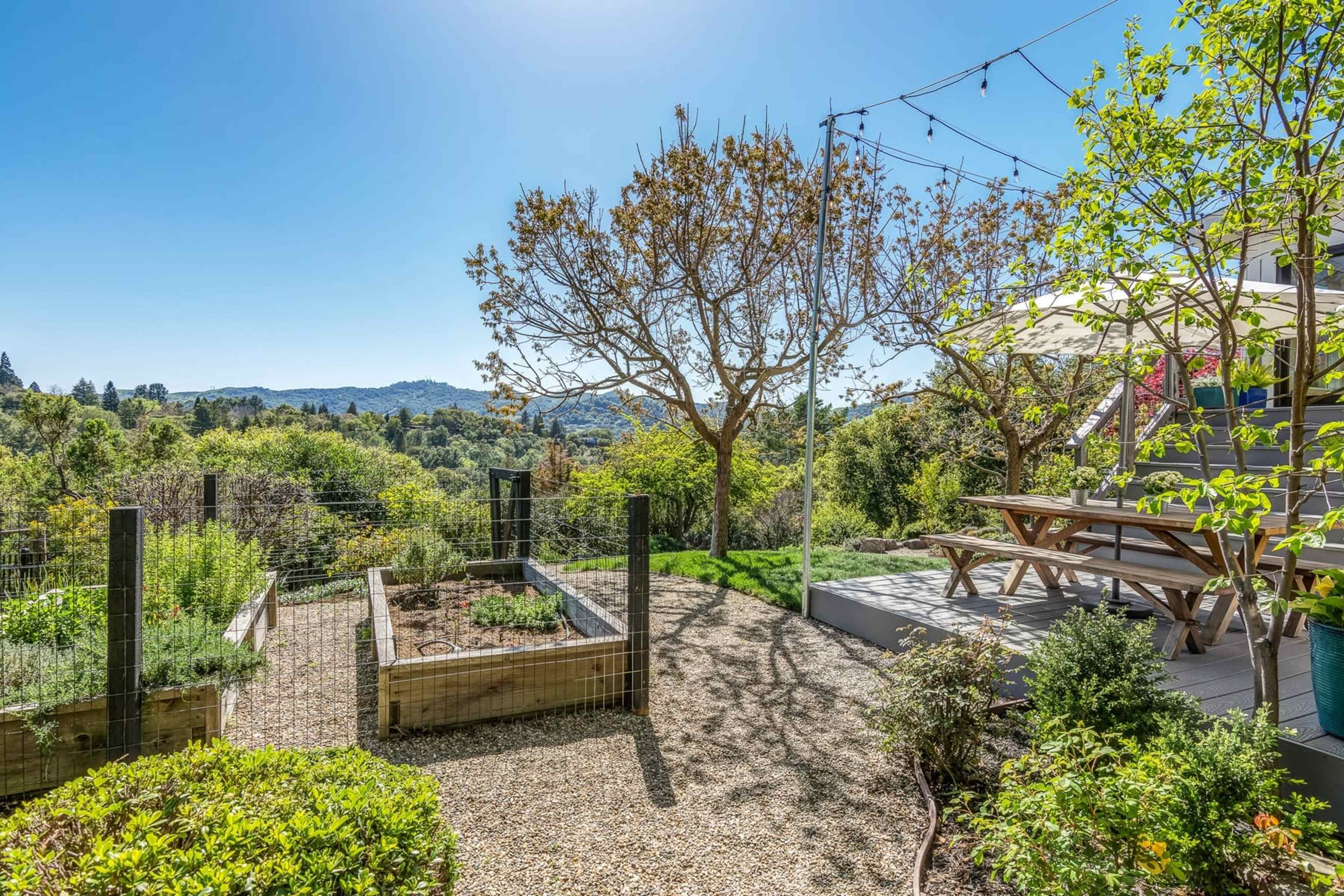 A landscaped backyard features a garden bed, a picnic table under a shade structure, and a view of rolling hills in the distance.