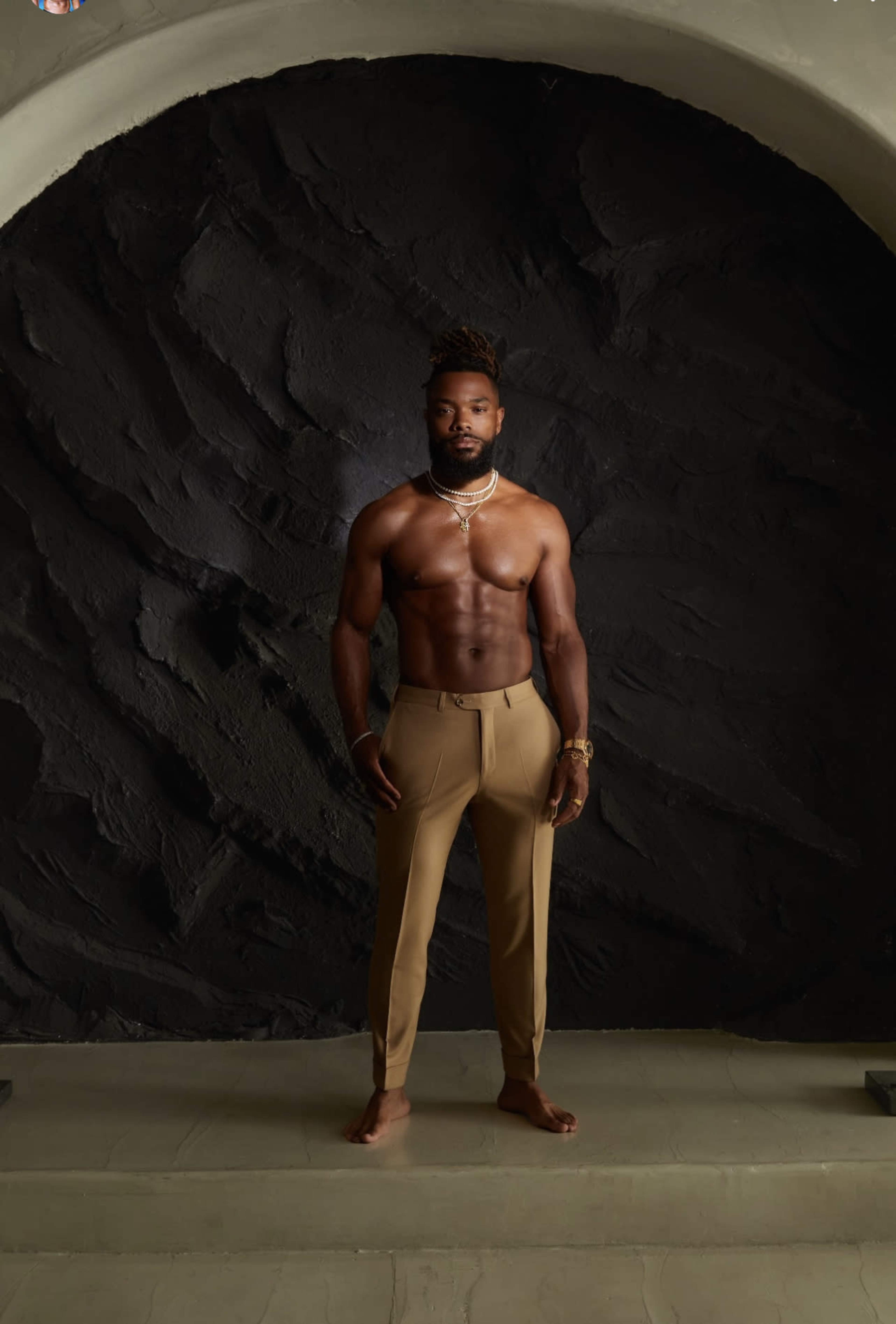 A man stands shirtless in tailored tan trousers against a dark textured backdrop.