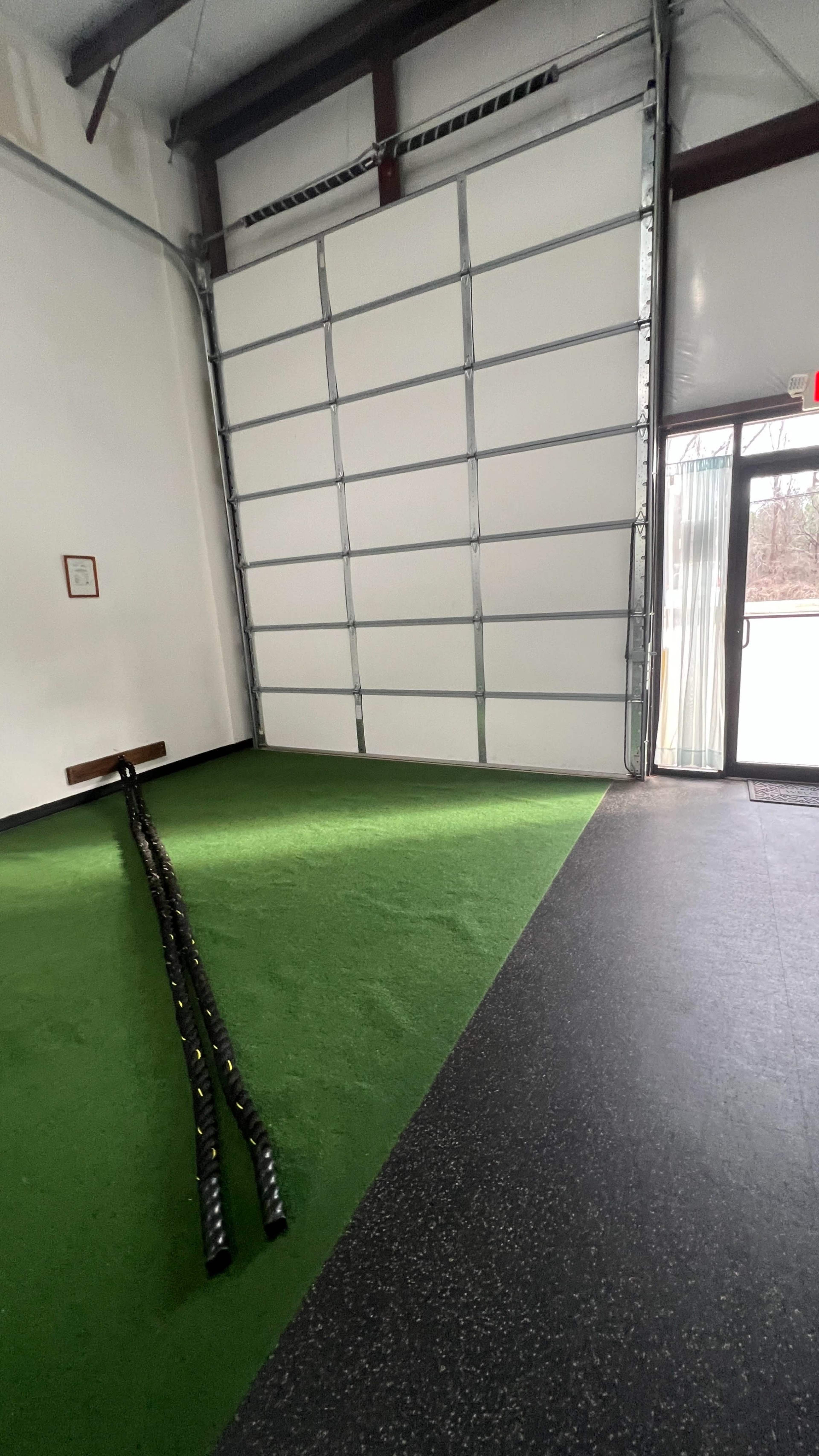 A gym or fitness facility with a green turf area, a large garage door on one side, and a doorway leading outside on the other.