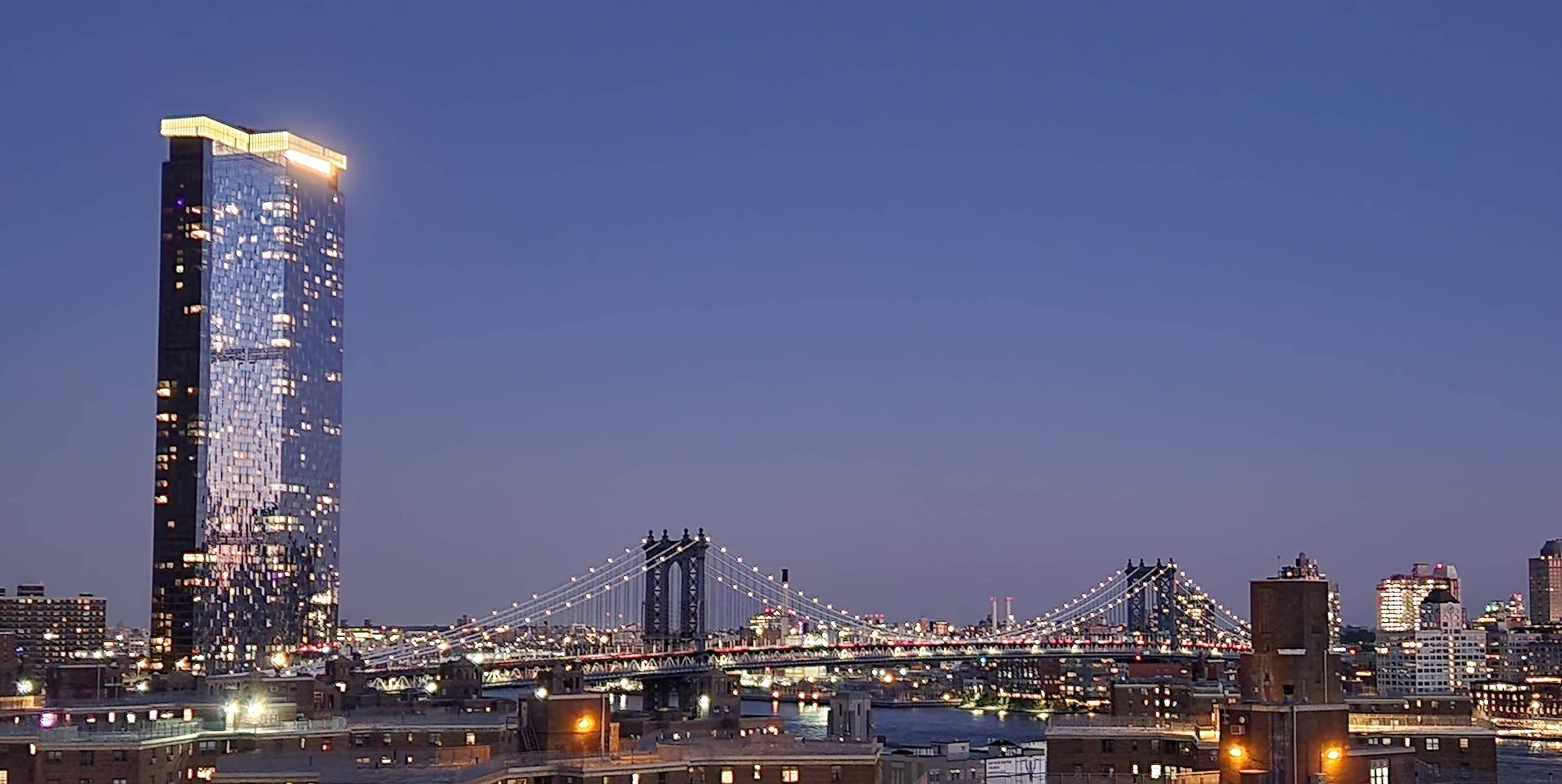 A night view of the Manhattan skyline, featuring a brightly lit skyscraper and the Brooklyn Bridge spanning the river.
