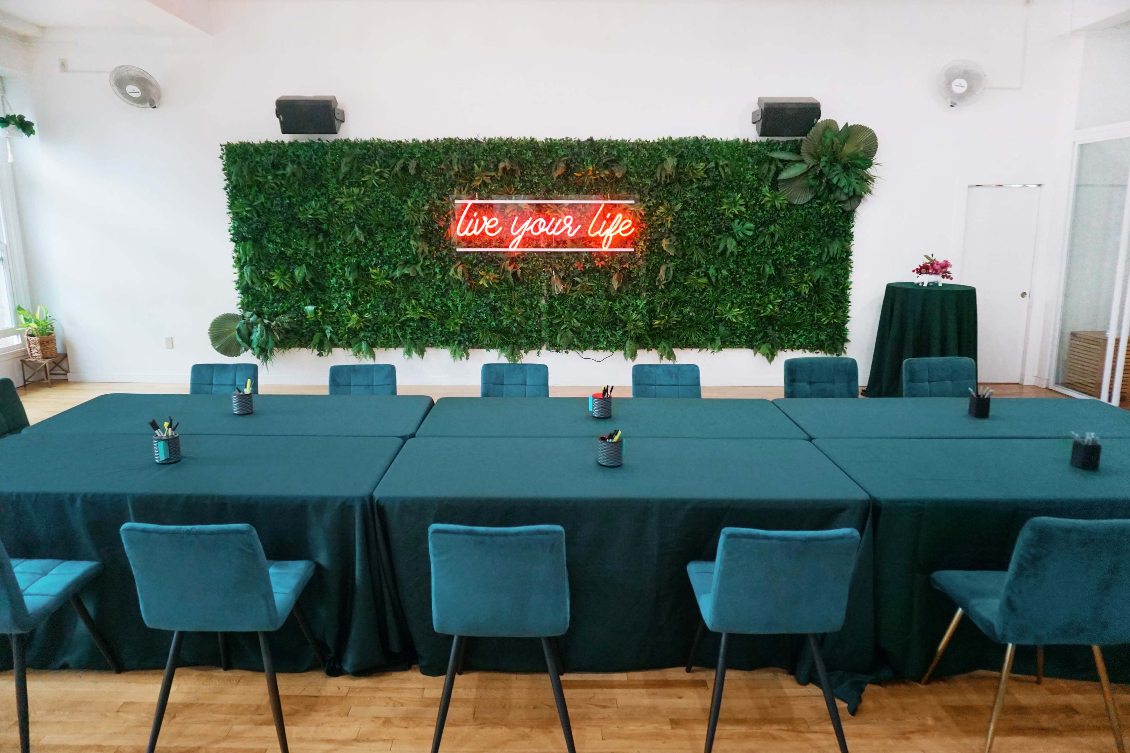 A large meeting space features a long table covered with dark green cloths and surrounded by blue chairs, with a greenery wall and a neon "live your life" sign in the background.