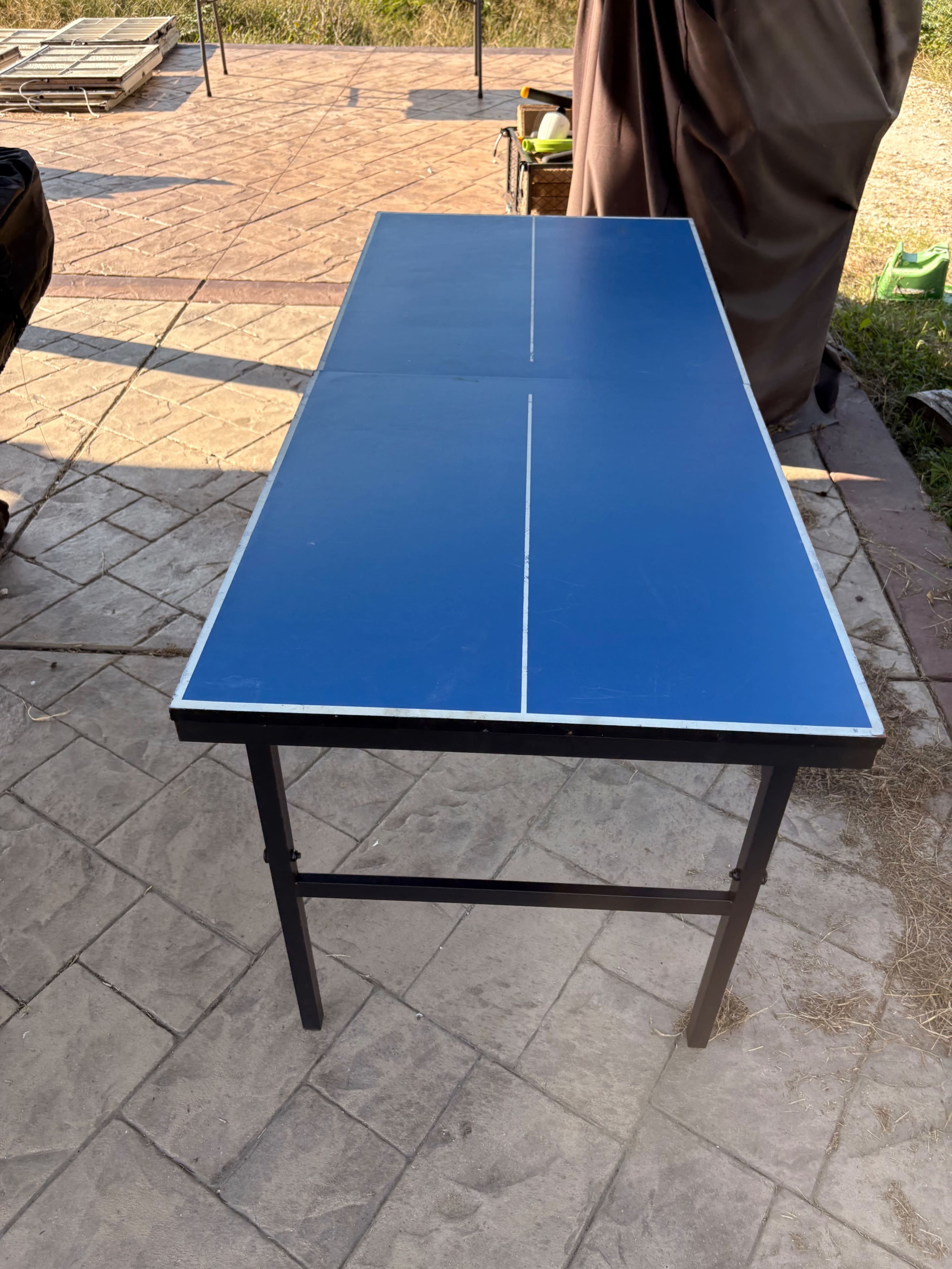 A blue ping pong table with a black metal frame is set up outdoors on a patterned stone surface.