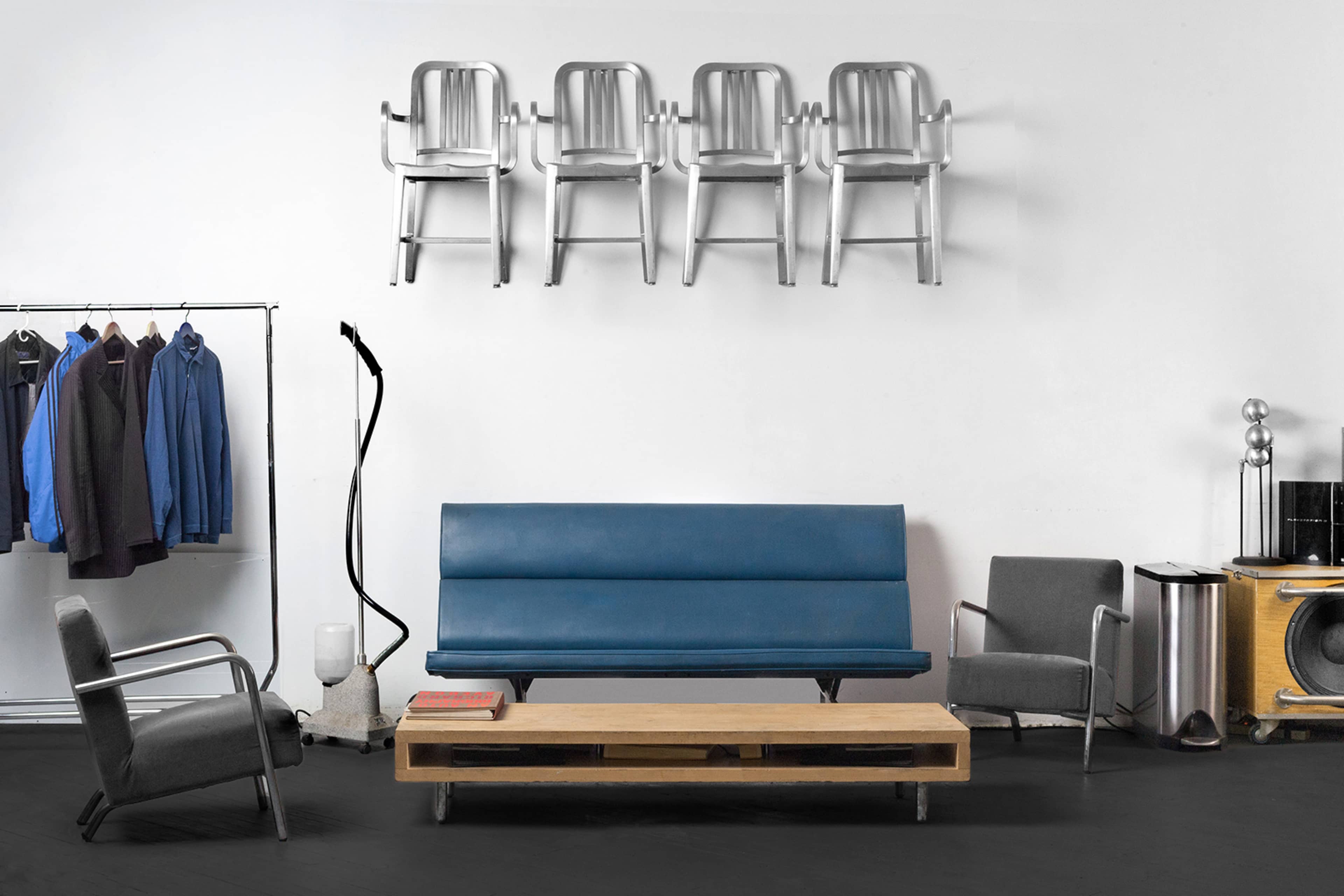 A modern living room with a blue sofa, a wooden coffee table, and several metal chairs hung on the wall, accompanied by clothing on a rack and a vintage speaker.
