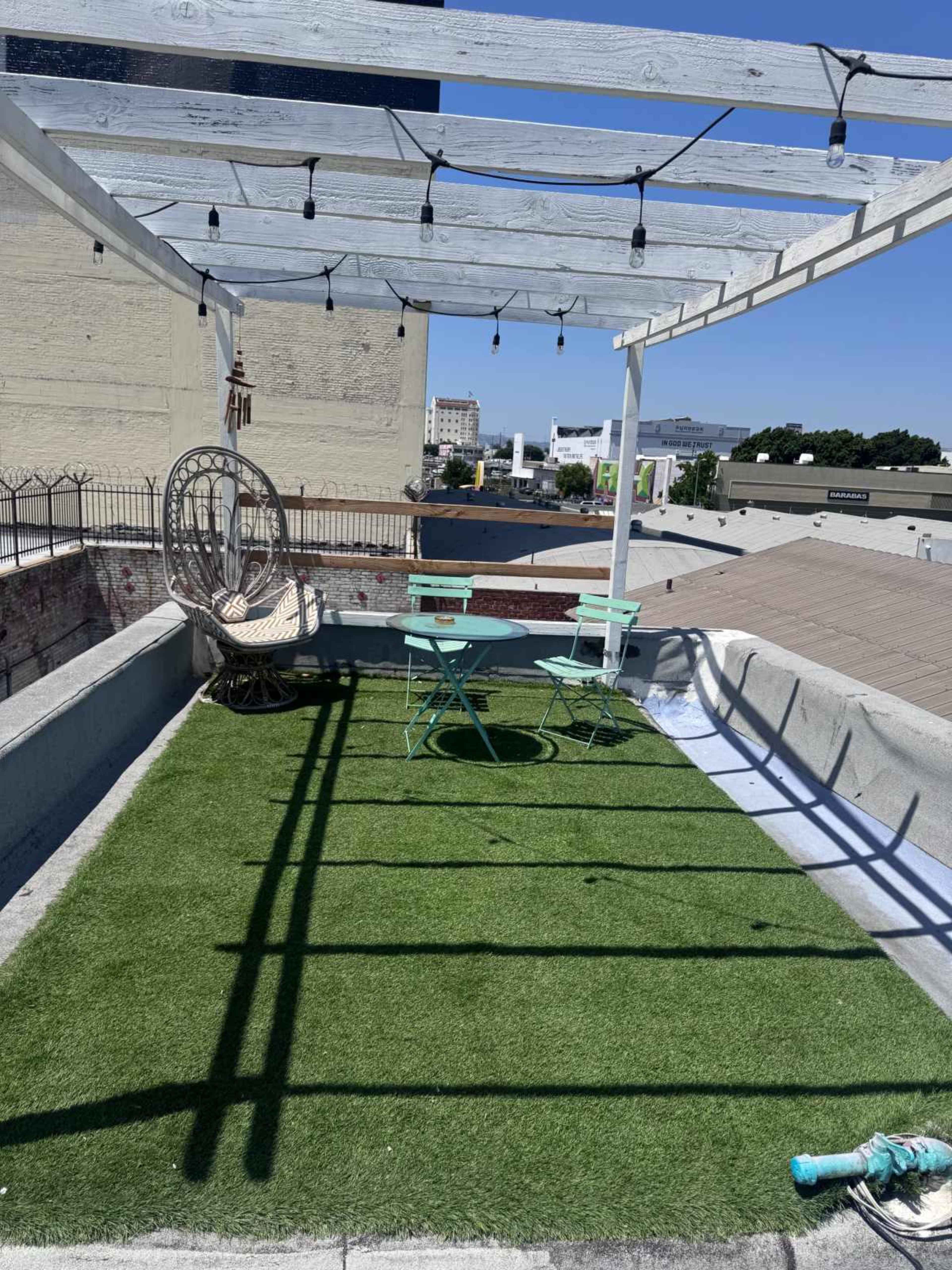A rooftop space features a grassy area with a small table and chairs, surrounded by a railing and string lights overhead.