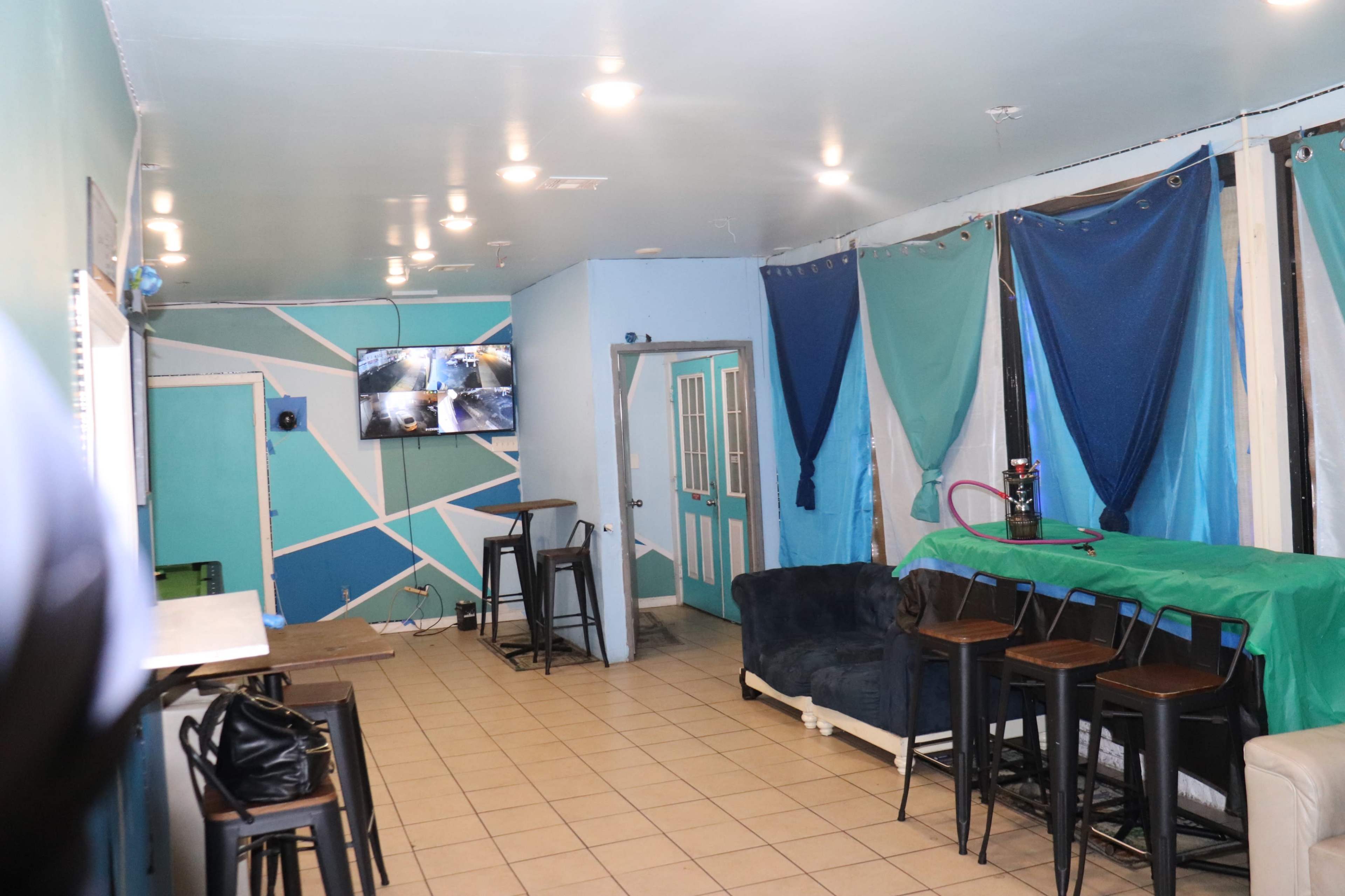 A lounge area features a couch, barstools, a television displaying security footage, and draped fabric in various colors.