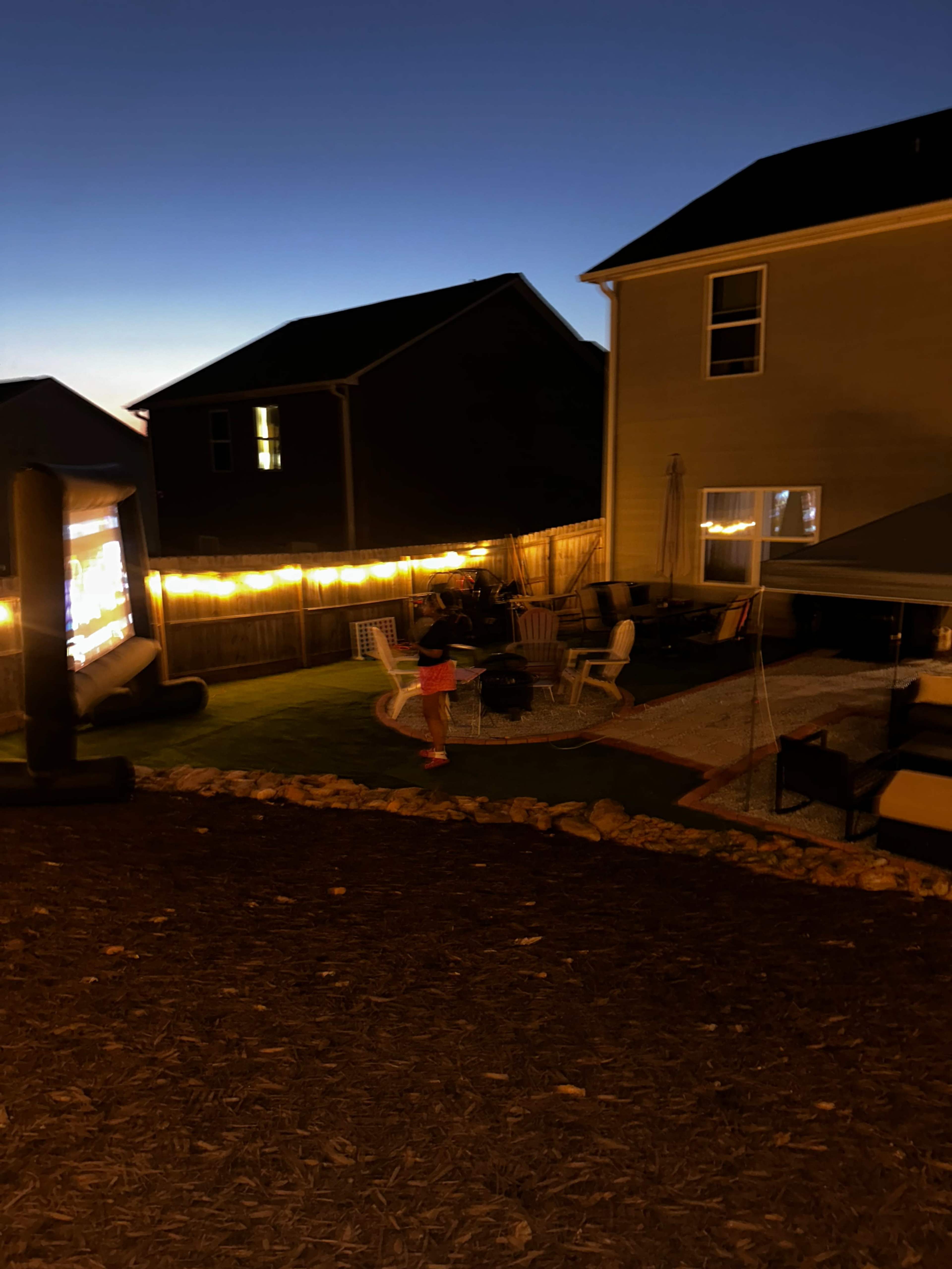 A backyard scene at dusk features a projection screen, seating areas with chairs, and string lights along a fence.