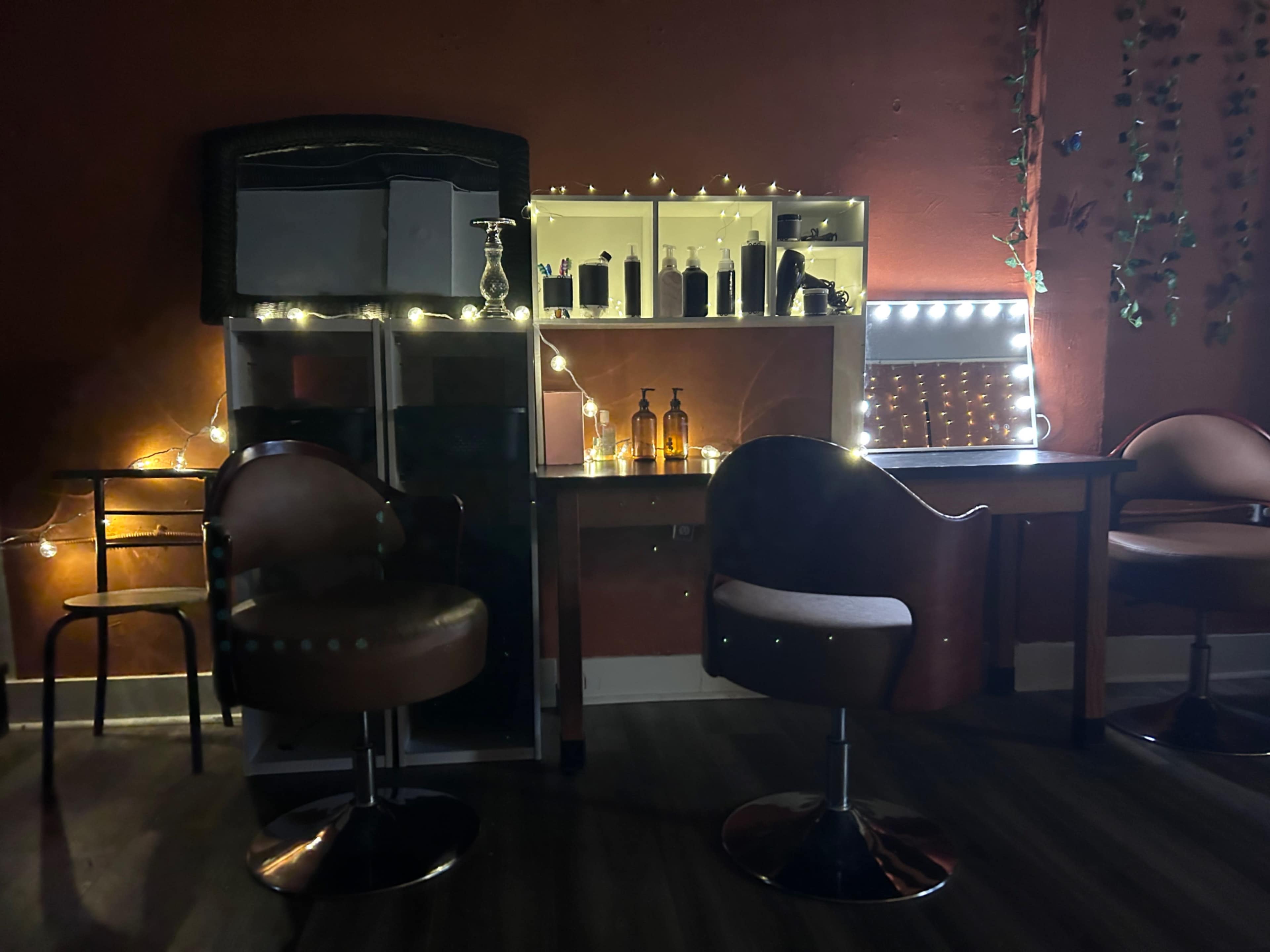 The image shows a dimly lit room featuring a mirrored vanity, shelves lined with bottles, and two chairs, enhanced by string lights along the walls.