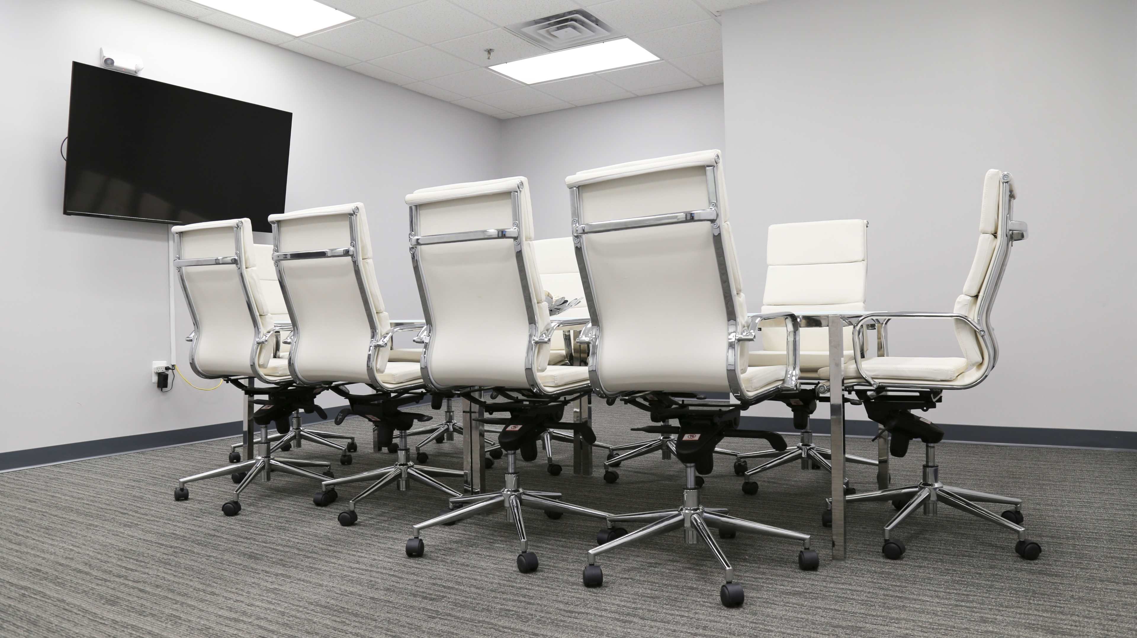 The image shows a conference room with a long table and several white office chairs arranged around it, and a wall-mounted TV at one end.