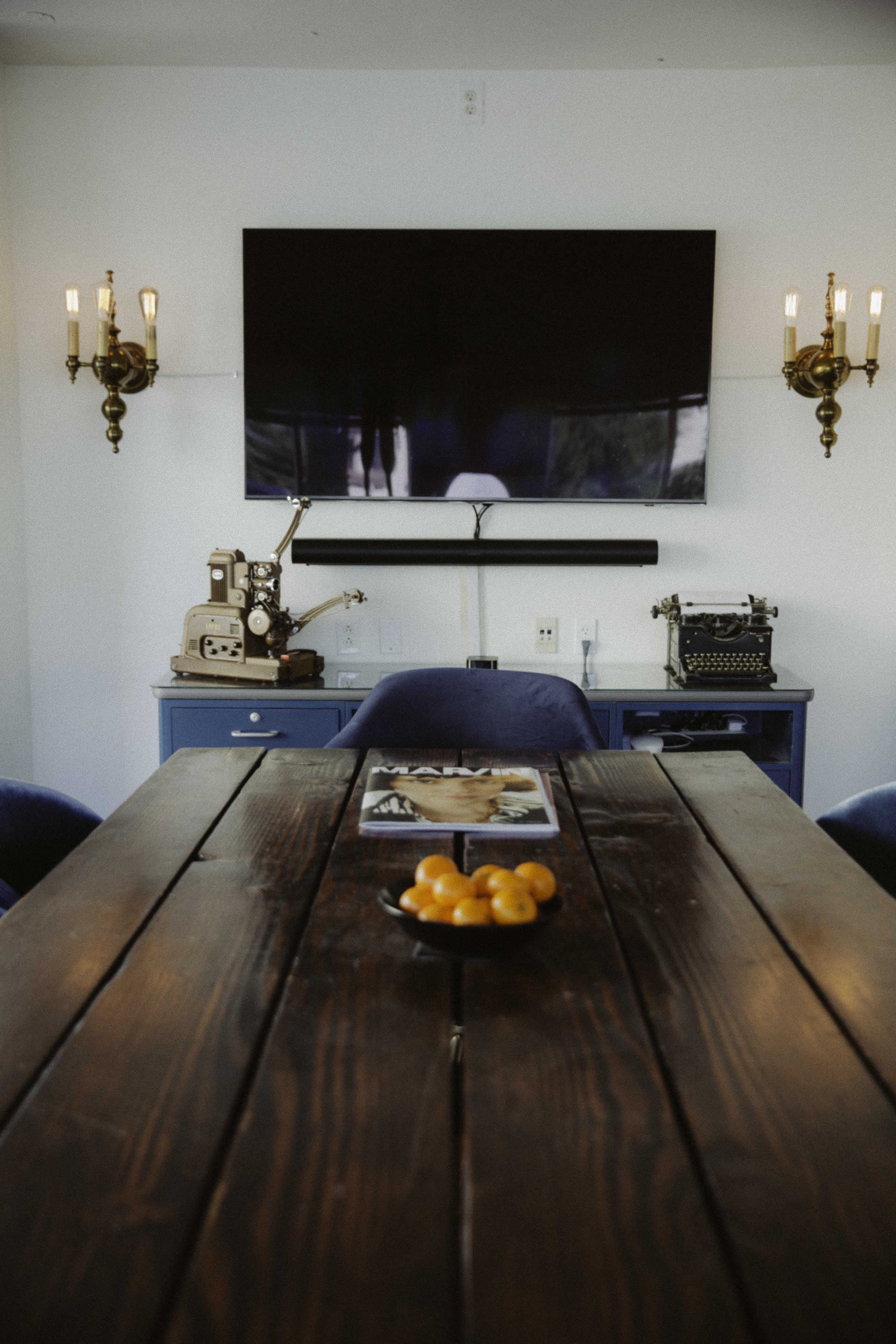 A wooden dining table with a bowl of fruit is centered in a room featuring a television mounted on the wall and vintage decor on a console.