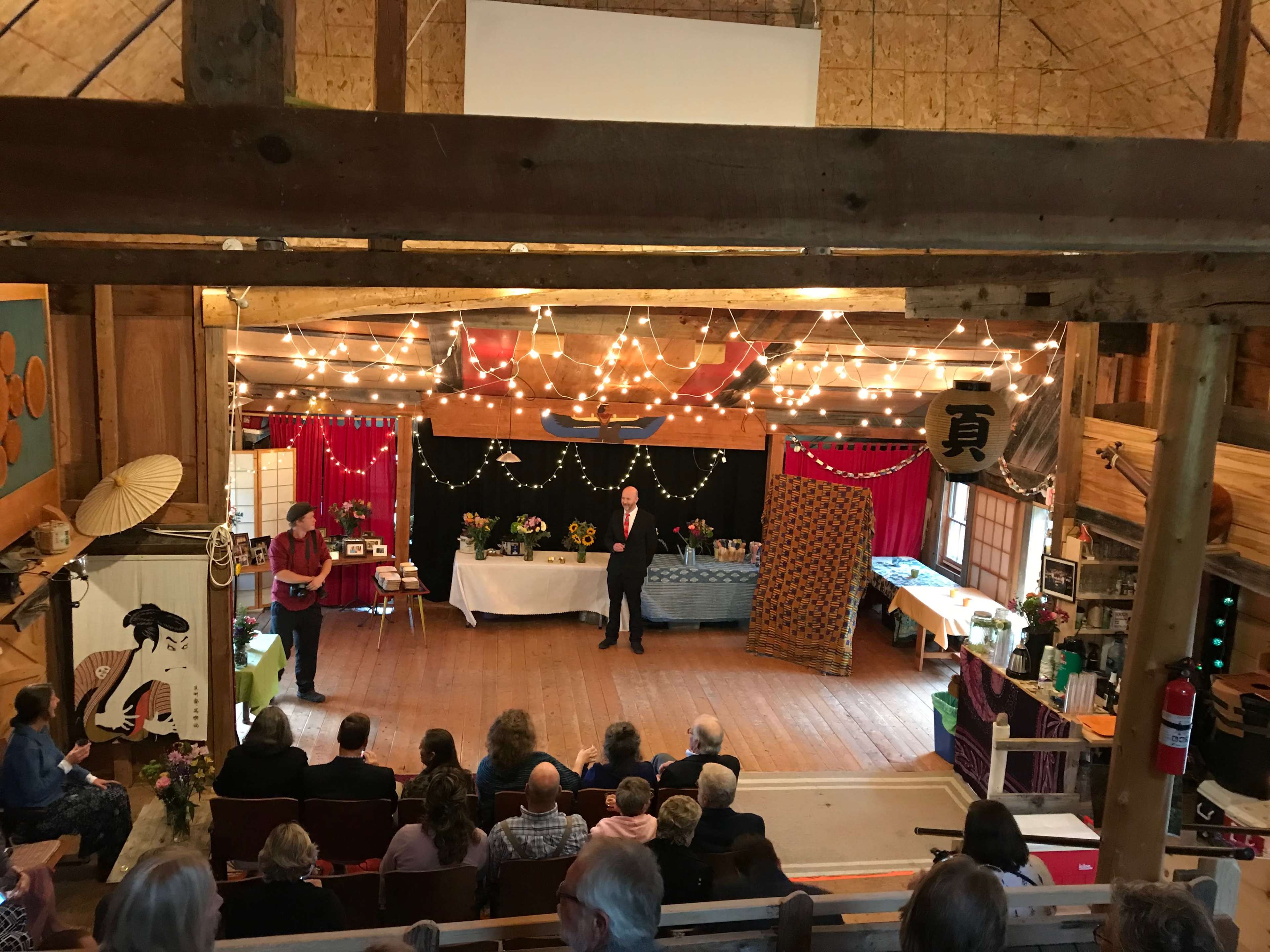A performance is taking place in a rustic hall decorated with lights, with an audience seated below and two speakers on stage.
