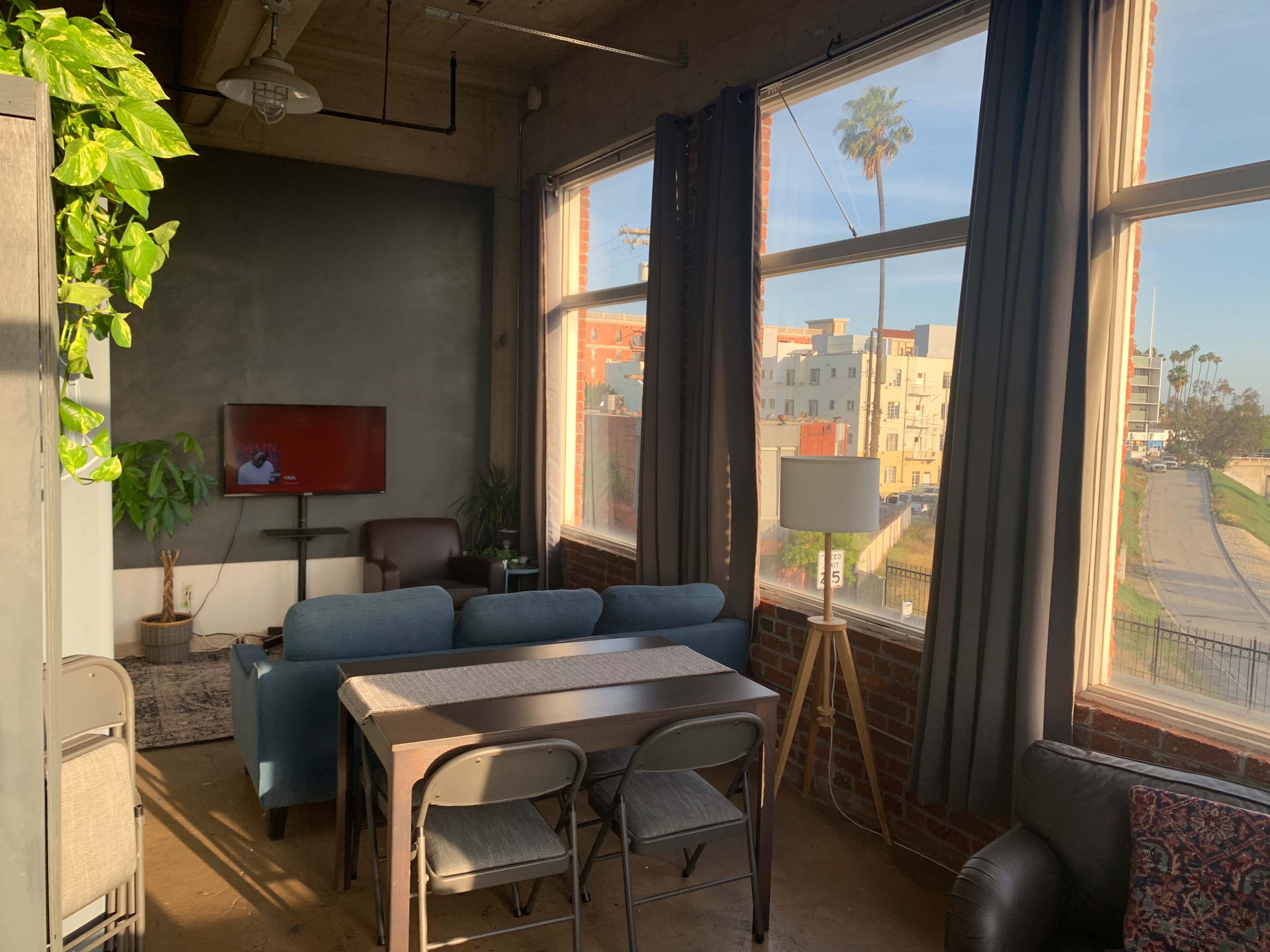 A living room with large windows allows natural light to illuminate a seating area, dining table, and television against a brick wall.