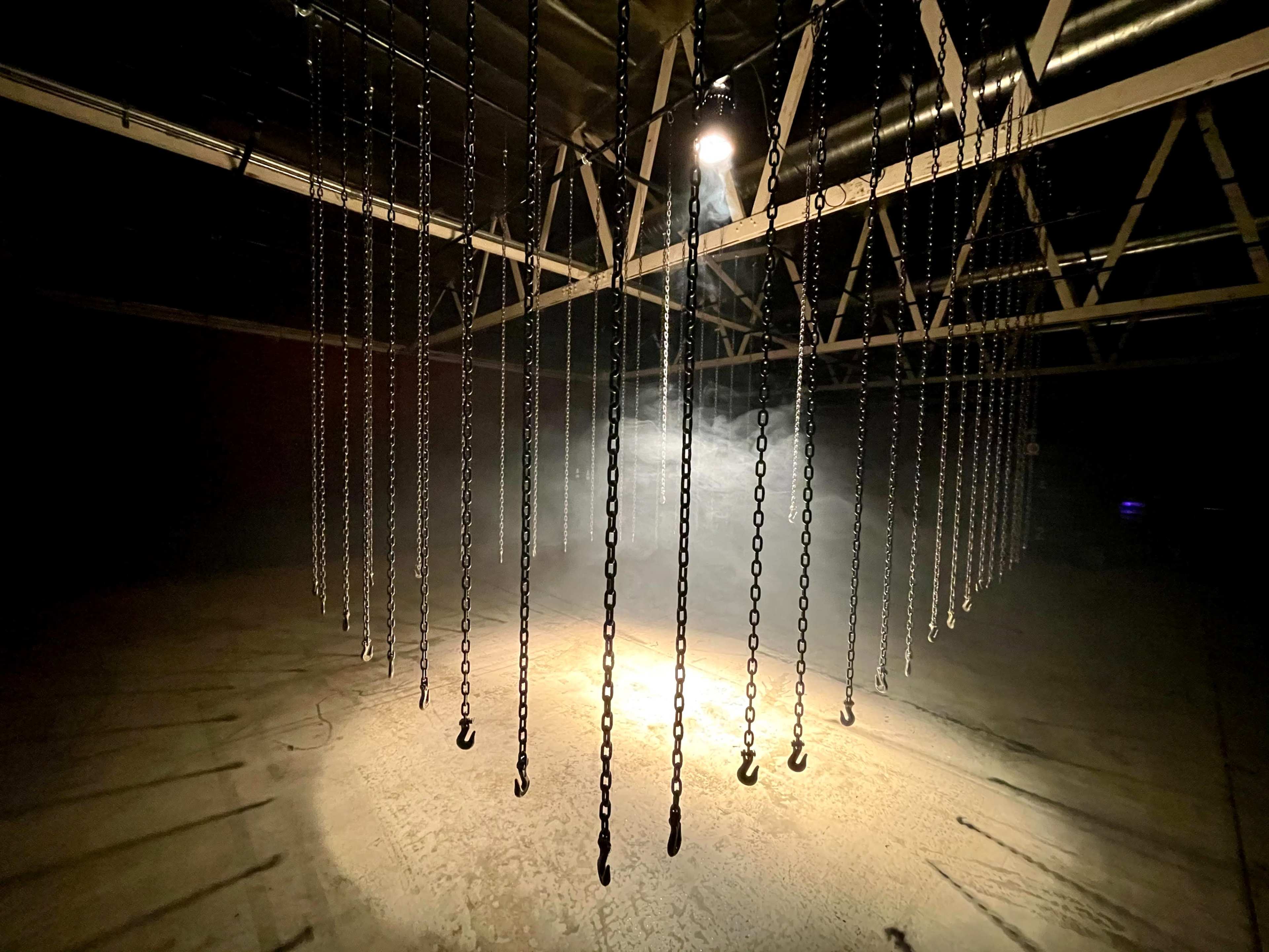 A dimly lit industrial space with chains hanging from a metal framework, illuminated by a single light source creating a spotlight effect on the ground.