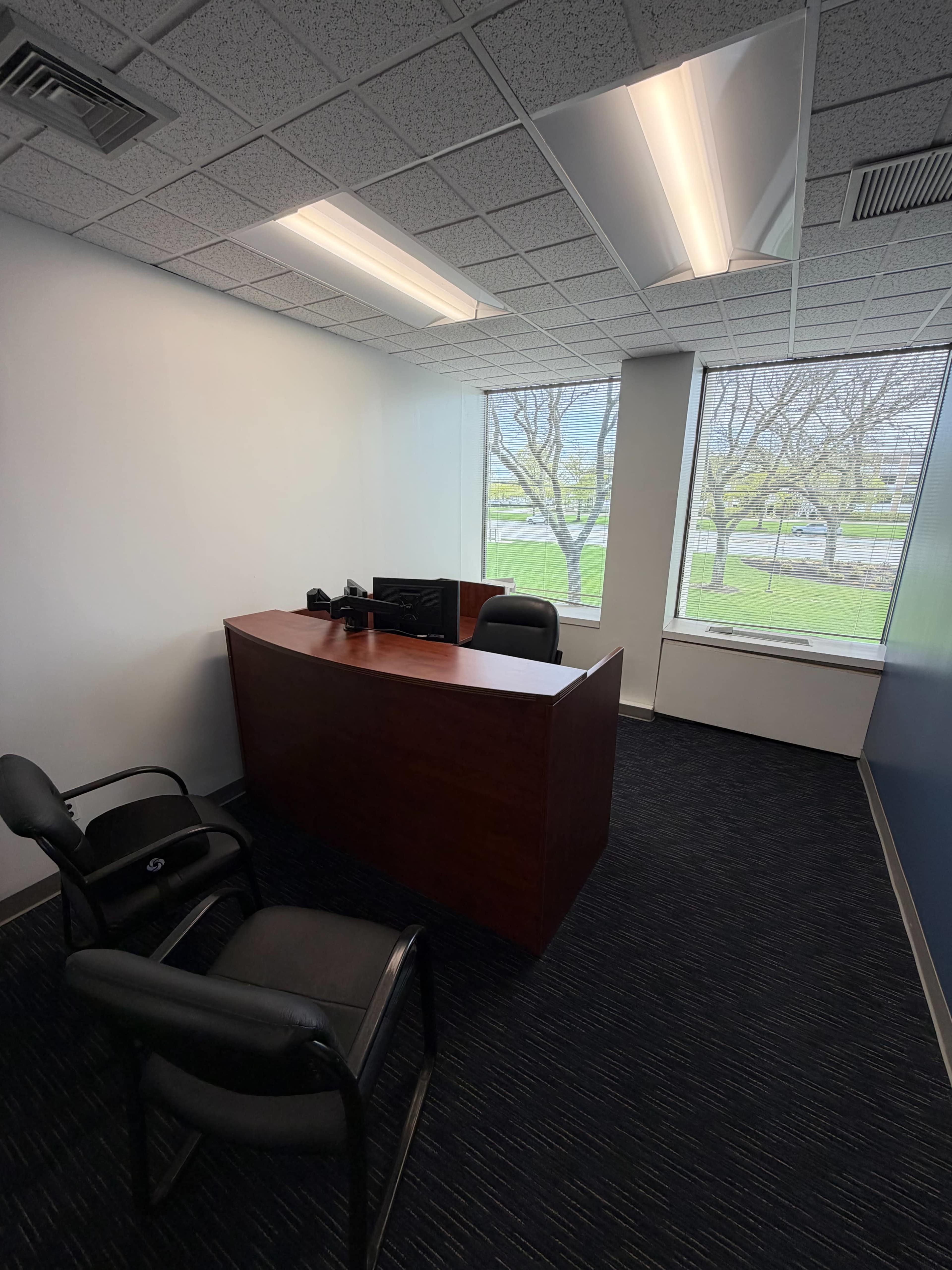 The image shows a small office with a wooden reception desk, two black chairs, and large windows overlooking a grassy area.