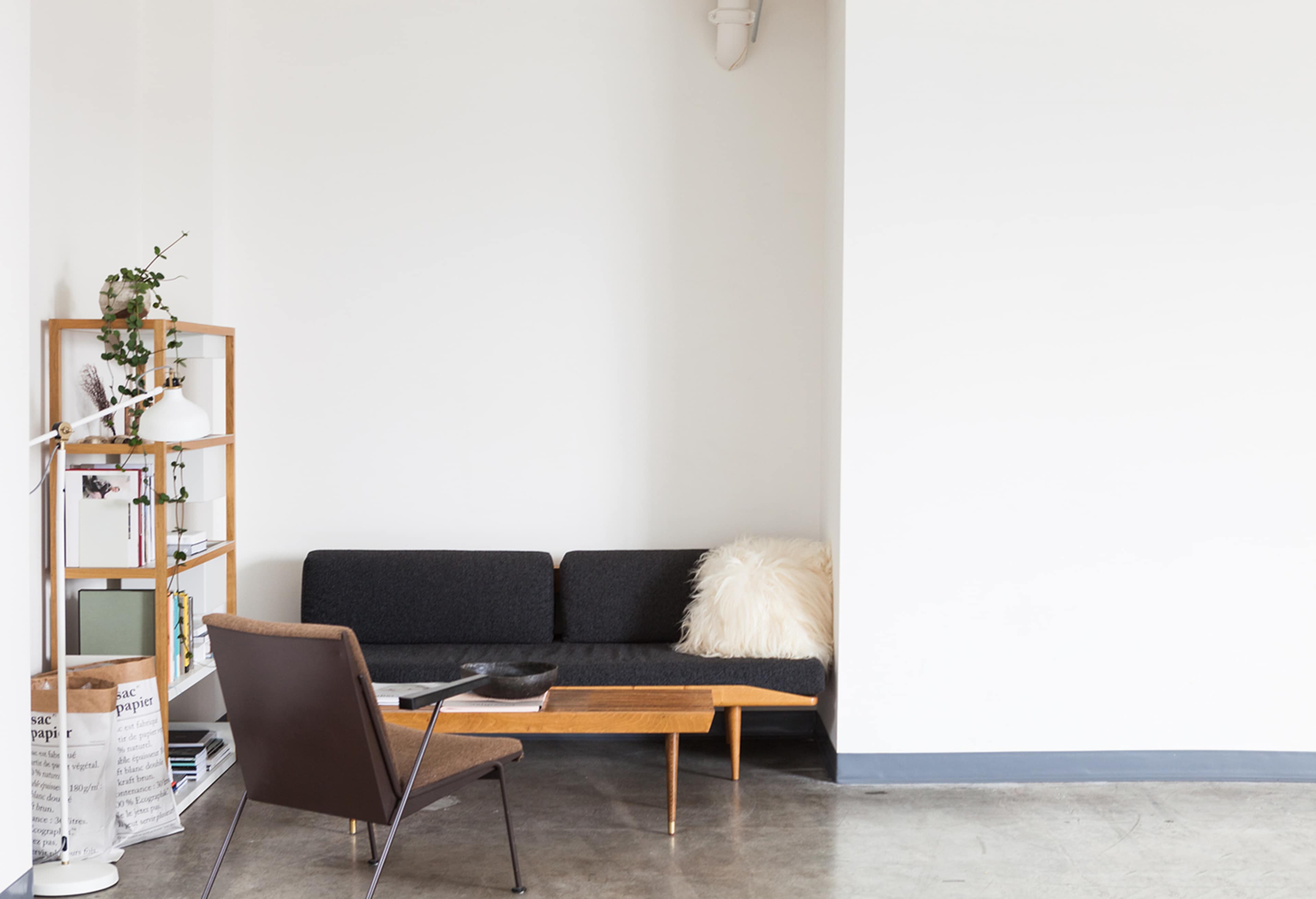 A minimalist living space with a dark sofa, a wooden coffee table, a chair, a floor lamp, and a bookshelf against a white wall.