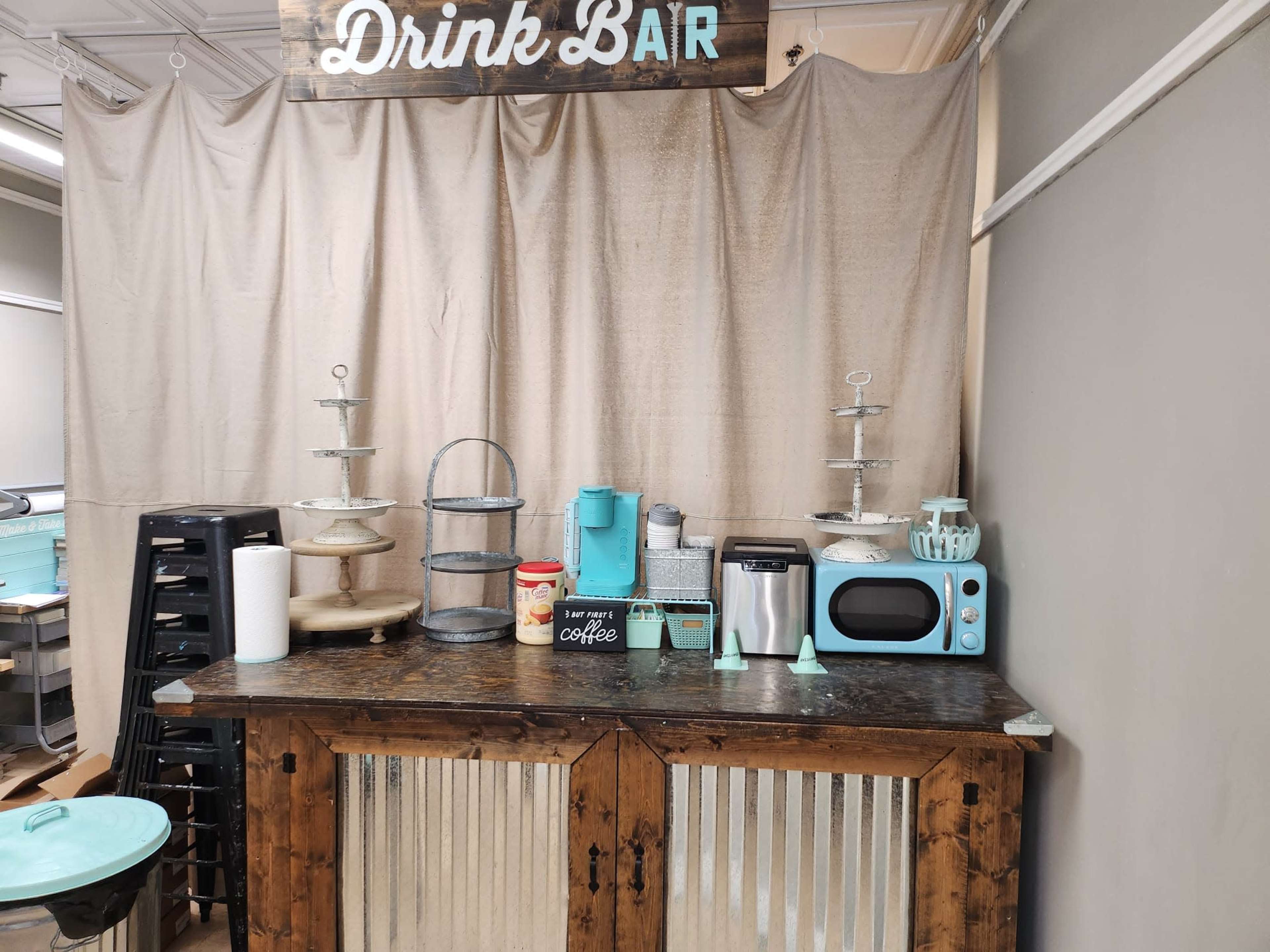 A drink bar setup featuring a wooden counter, various kitchen appliances, and decorative containers against a backdrop of a beige curtain.