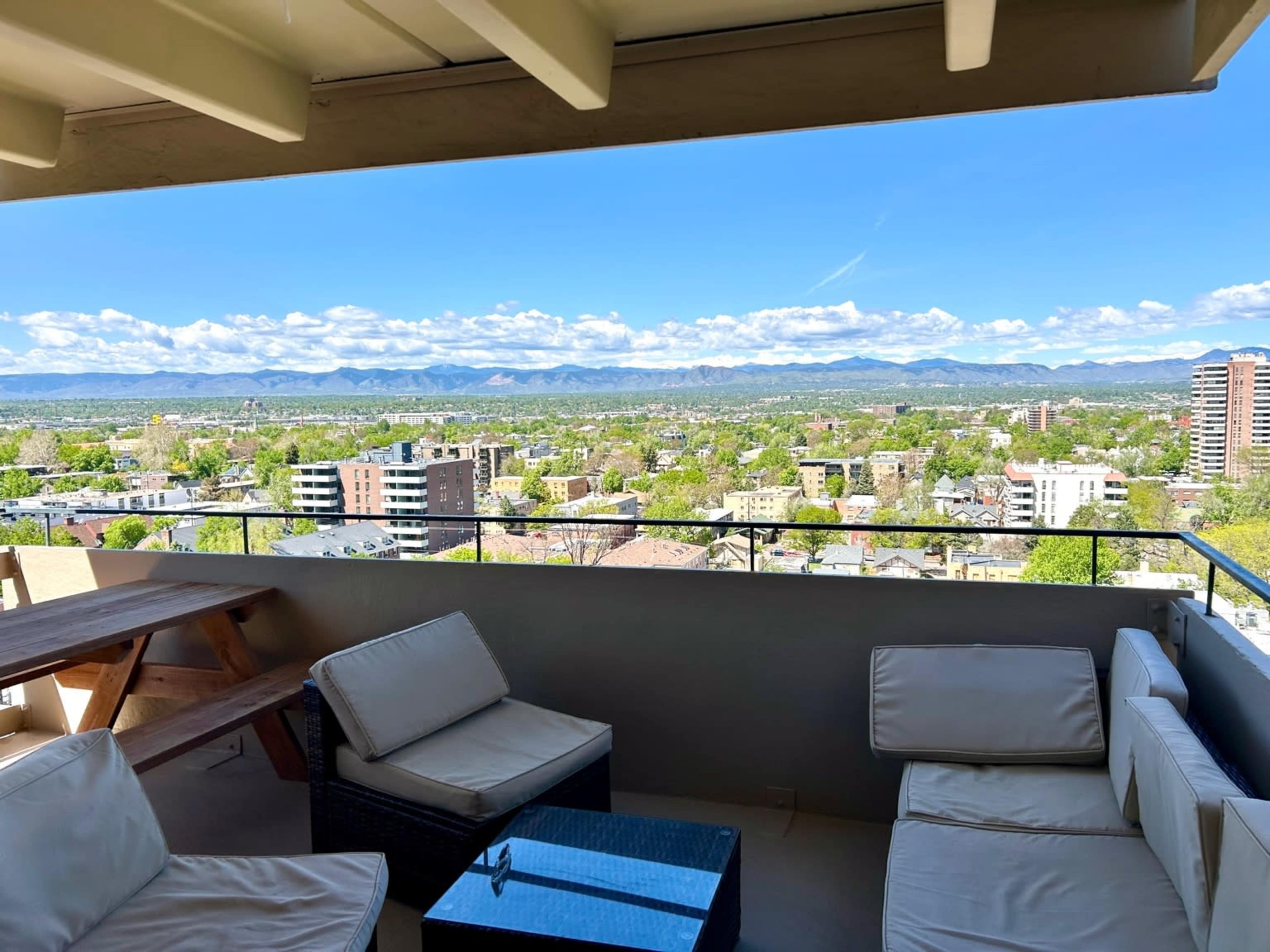 The image shows a balcony with modern seating and a glass table, overlooking a cityscape and mountainous horizon under a blue sky.
