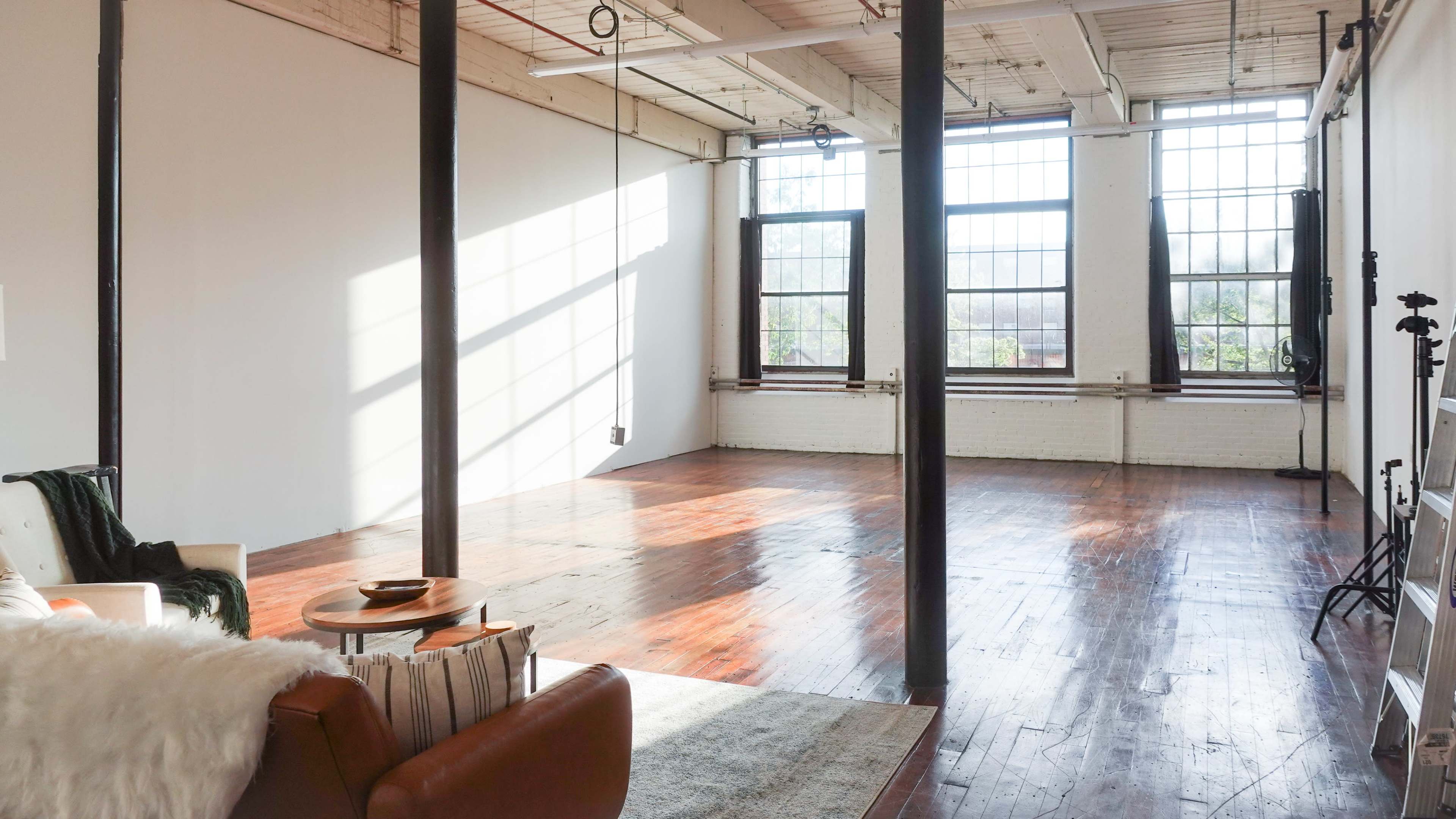 A spacious, empty studio with large windows that let in natural light, wooden floors, and minimal furnishings.
