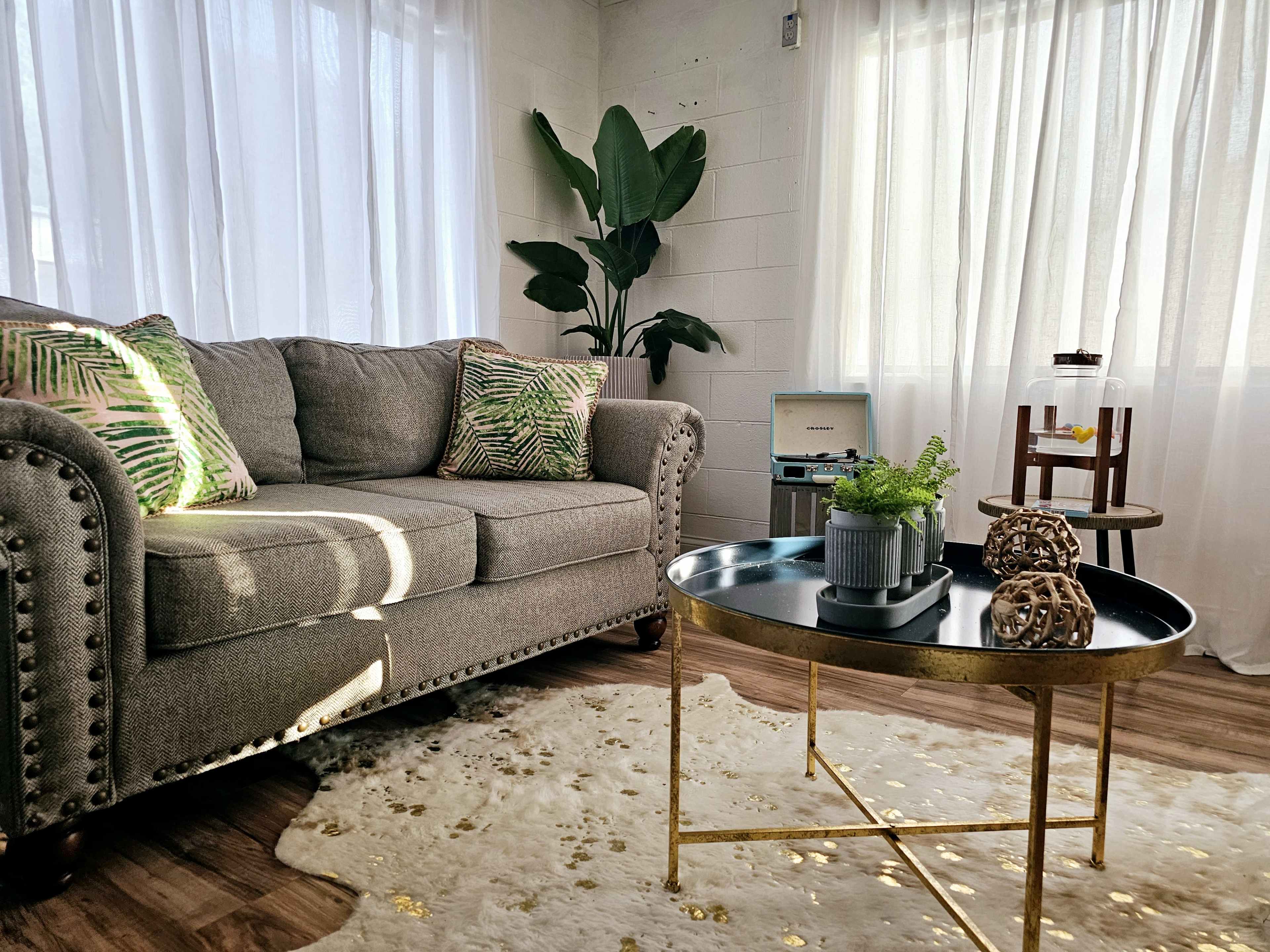 A cozy living room featuring a gray couch with patterned pillows, a coffee table with decorative items, and a small plant next to a large window with sheer curtains.