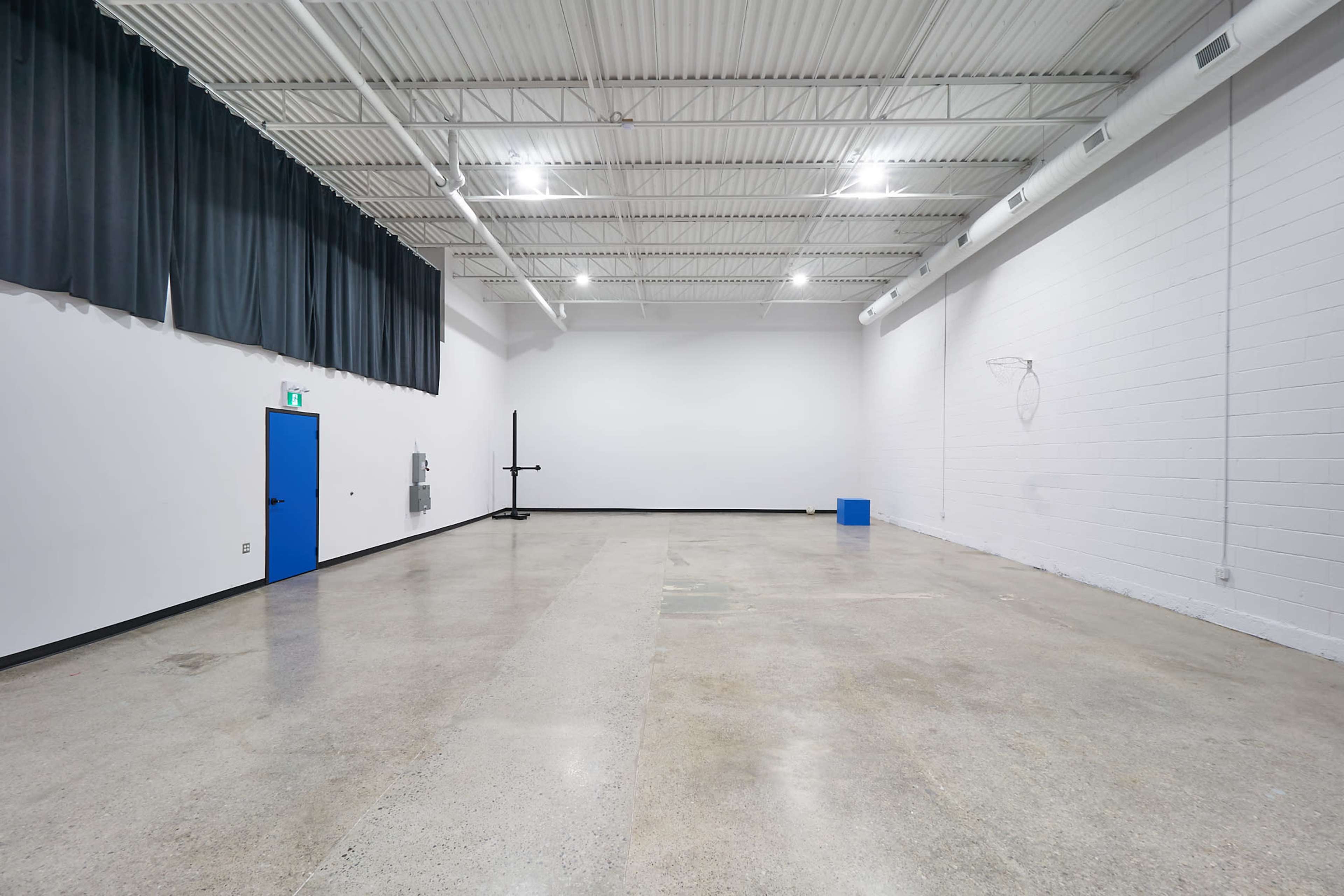 A large, empty gymnasium with polished concrete floors, high ceilings, and a blue door on one wall.