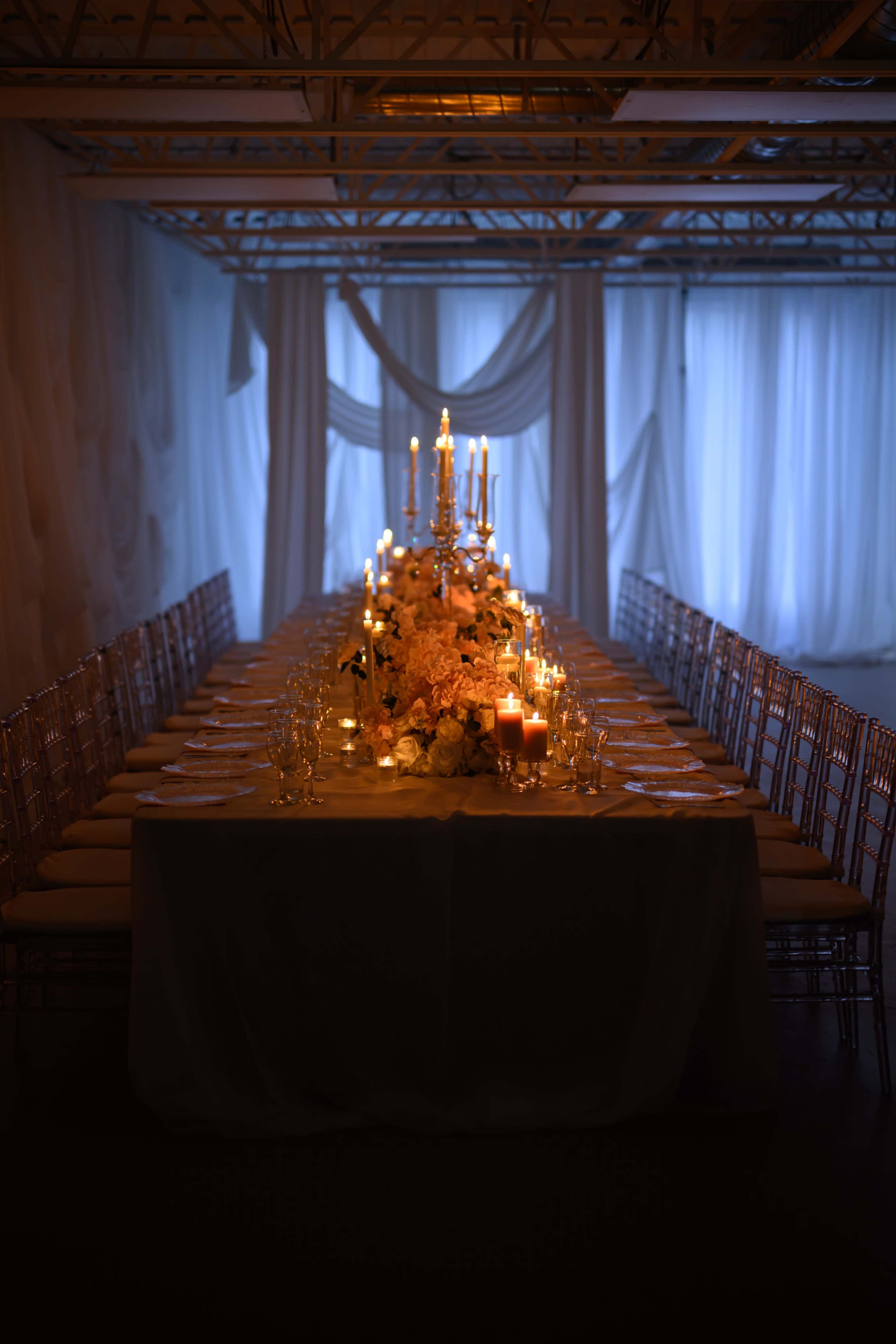 A long dining table is elegantly set with candles and floral arrangements under soft, dim lighting in a room with sheer drapes.