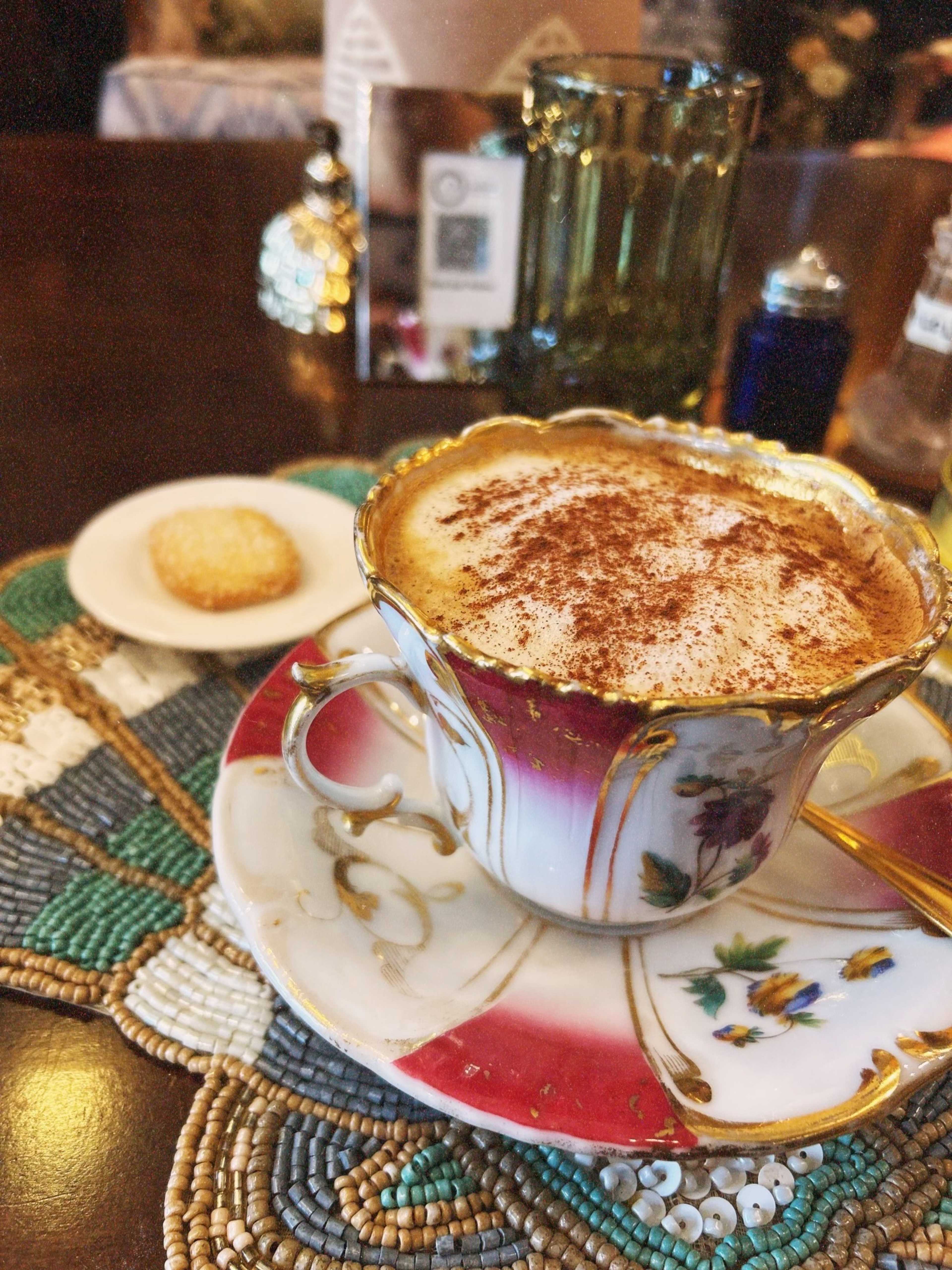 A beautifully designed cup of coffee topped with cocoa sits on a decorative coasters next to a small cookie.