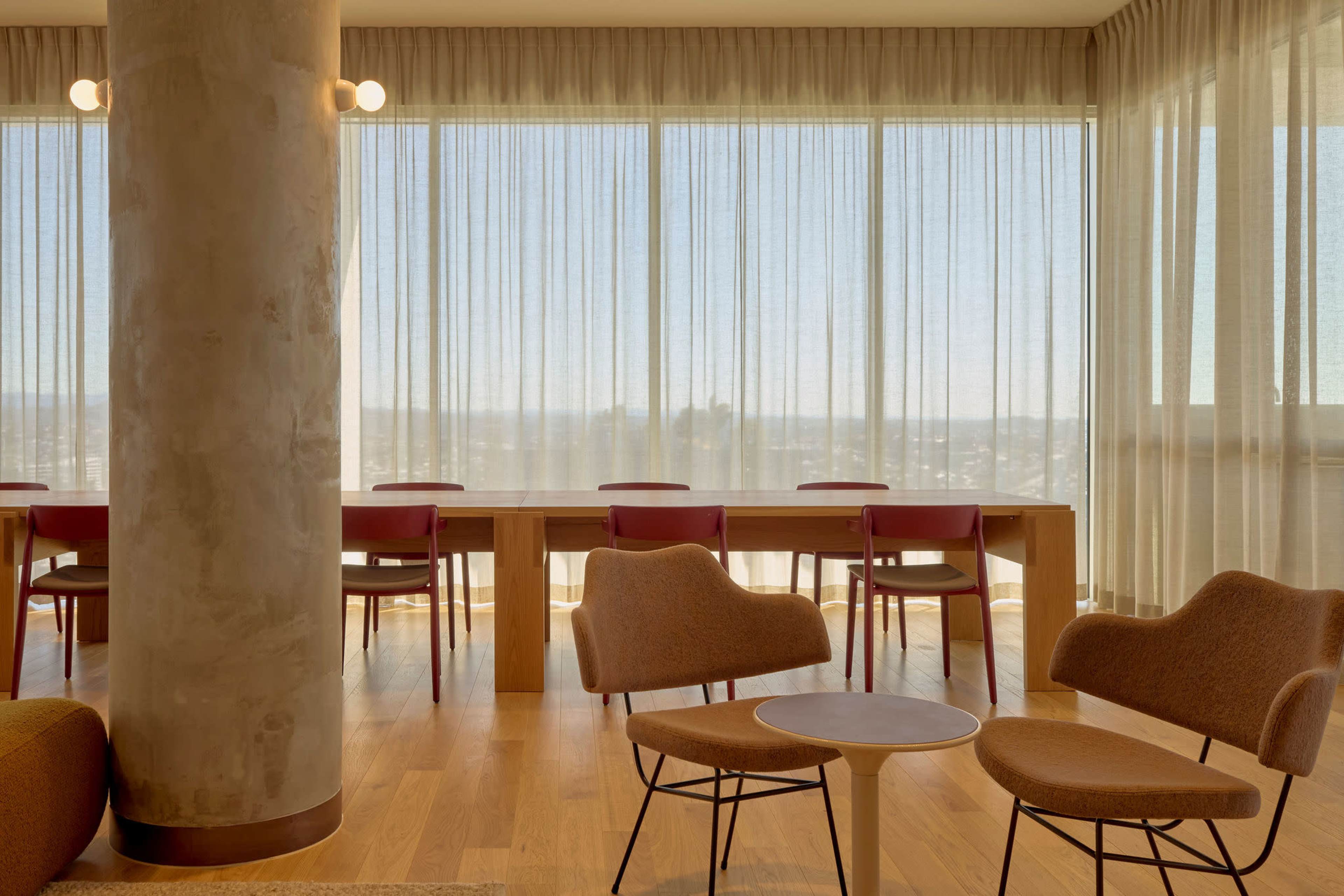 The image shows a modern interior space with a long wooden table surrounded by chairs, large windows with sheer curtains, and an inviting seating area.